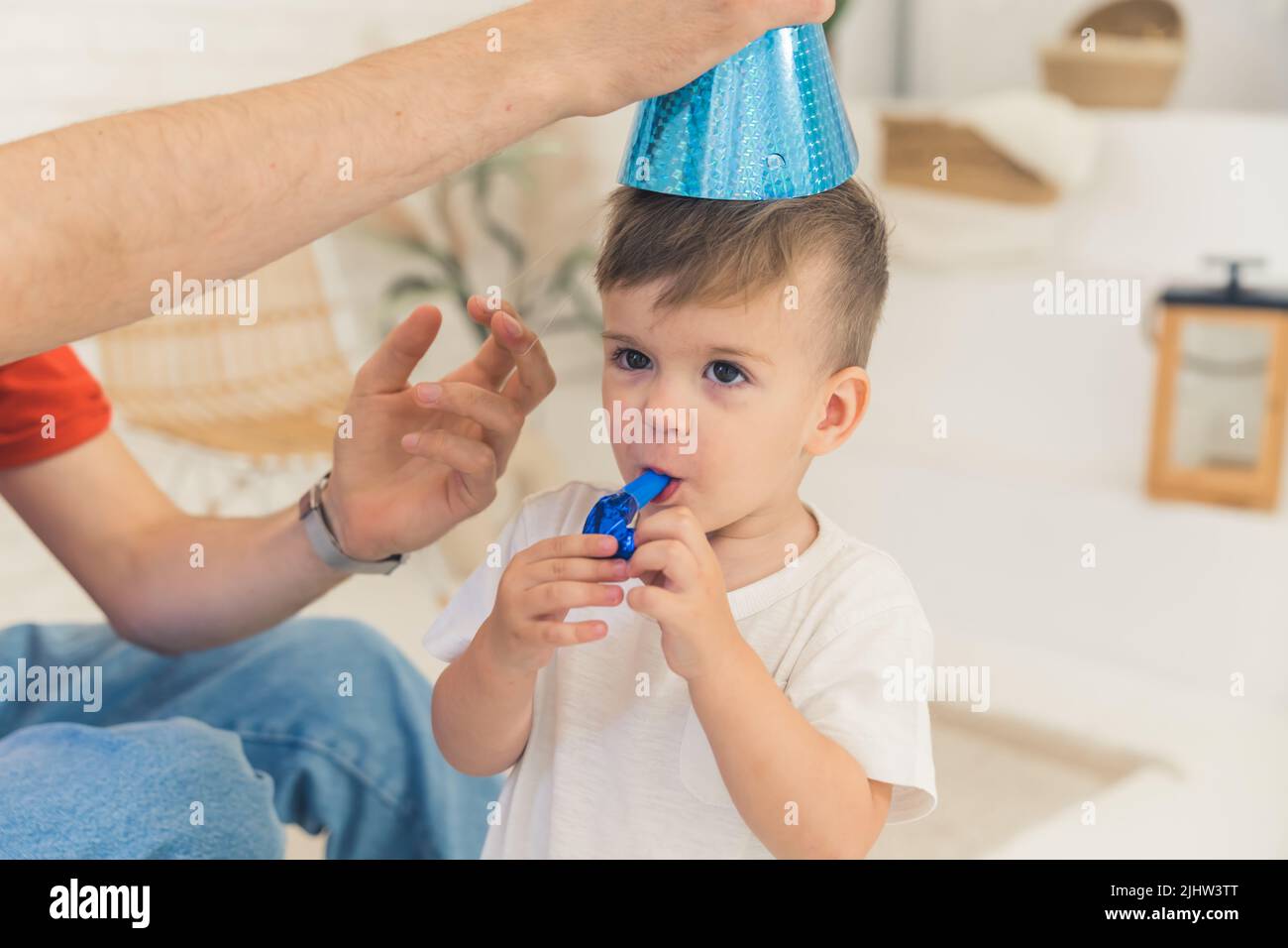 Liebenswert Kleinkind Junge mit einem funkelnden Party-Tasse auf dem Kopf versucht, ein Party-Horn zum ersten Mal blasen. Konzept zur Entwicklung von Kindern. Hochwertige Fotos Stockfoto