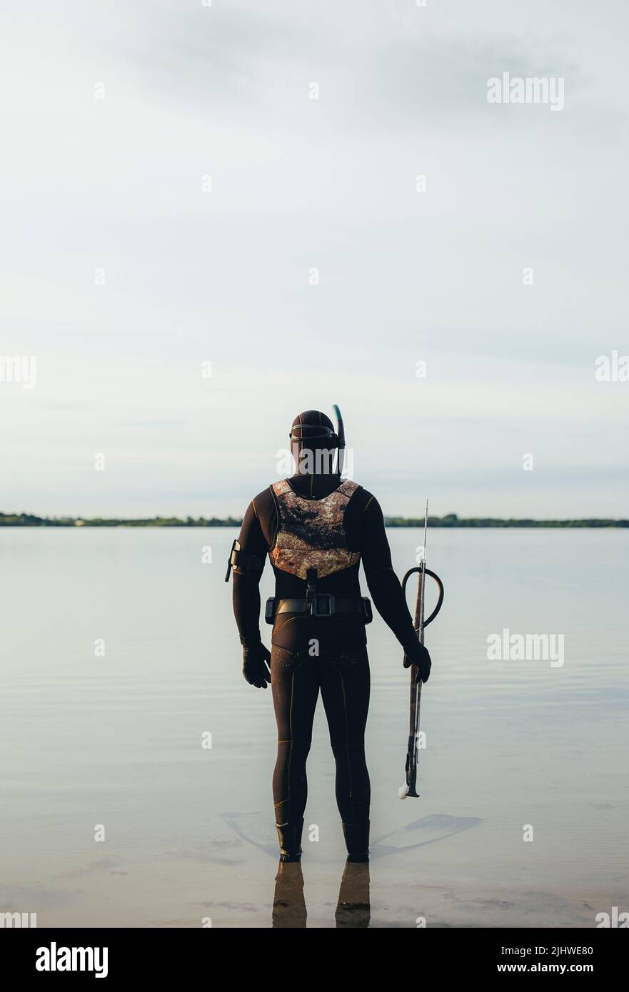 Mann, der eine Pistole in der Hand hält und im Meerwasser steht. Rückansicht eines Mannes in einem Neoprenanzug, der sich bereit gemacht hat, unter dem Wasser zu Speerfischen. Stockfoto
