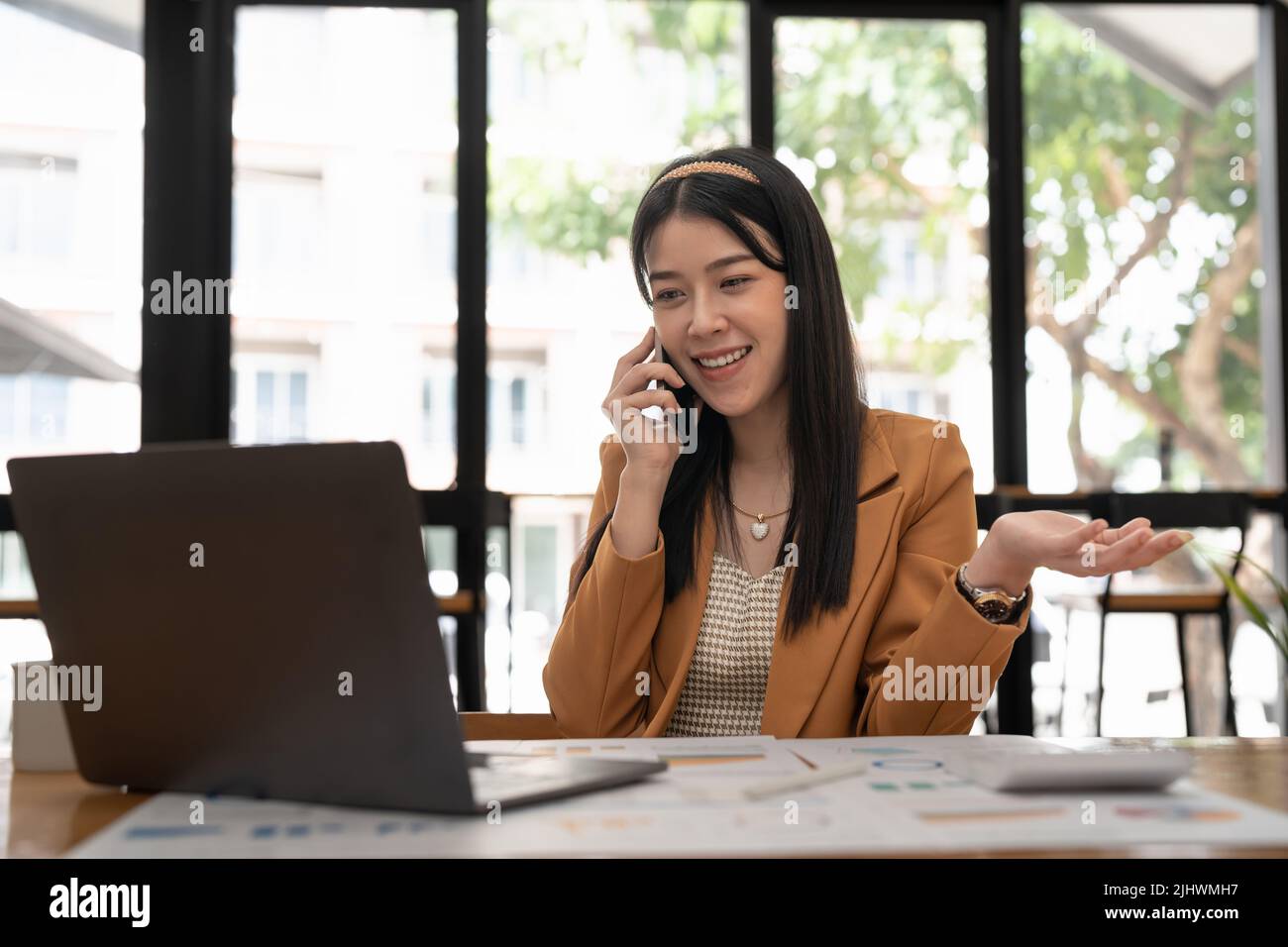 Fröhliche asiatische Angestellte, die mit Computer arbeitet und auf dem Mobiltelefon spricht. Zufriedene Geschäftsfrau, die einen Anruf mit dem Firmenkunden hält und darüber spricht Stockfoto