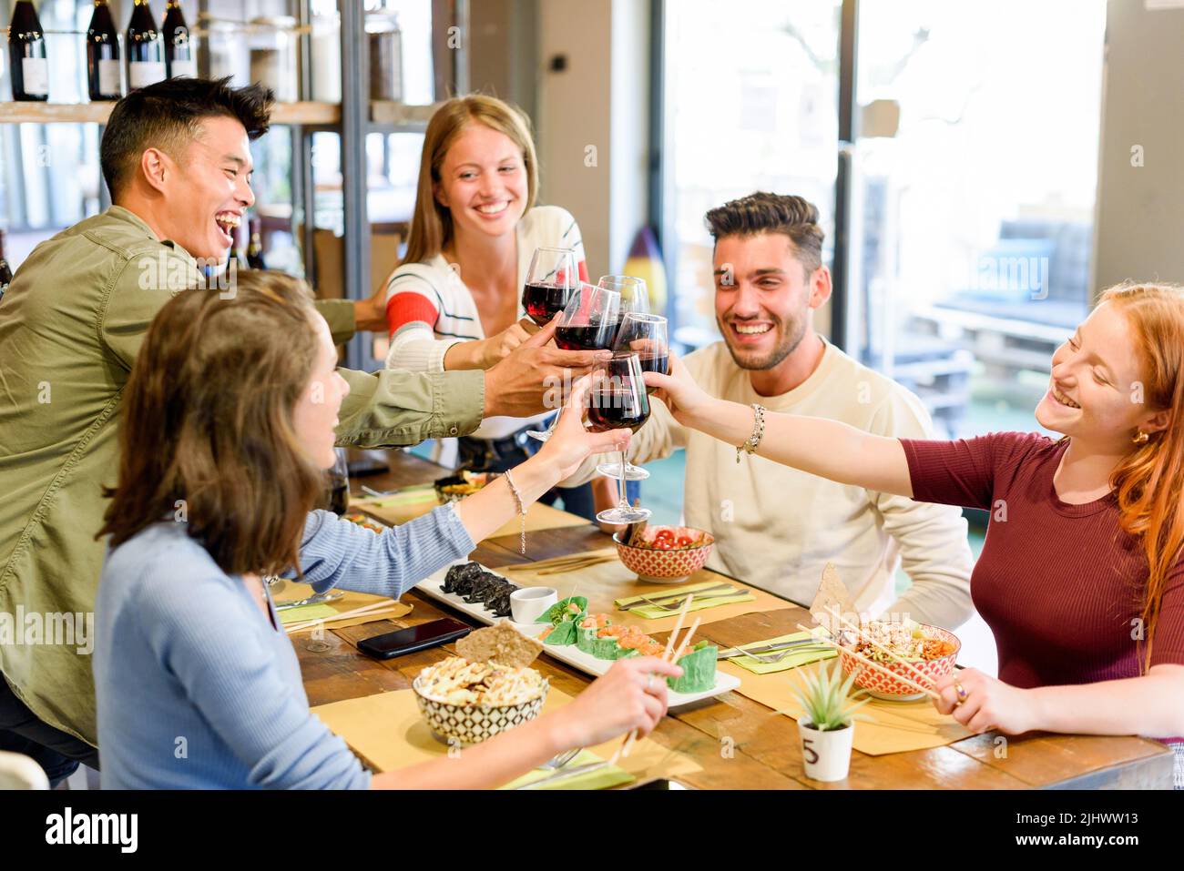 Eine Gruppe fröhlicher multirassischer Freunde, die sich gegenseitig angucken und mit Rotwein klirrende Weingläser anklirren, während sie gemeinsam am Tisch mit Essen feiern Stockfoto