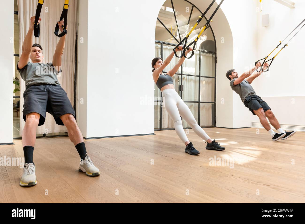 Vollkörper starker Menschen in der aktiven Trainingsbekleidung, die während des Funktionstrainings in einem modernen, leichten Fitnessstudio Stretching-Übungen mit TRX-Trägern machen Stockfoto