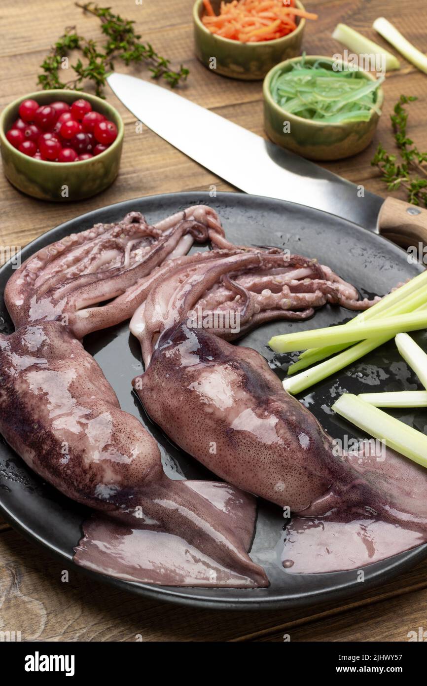 Roher Tintenfisch mit Tentakeln auf schwarzem Teller. Karotten, Algen und Beeren in Schüsseln. Draufsicht. Holzhintergrund Stockfoto