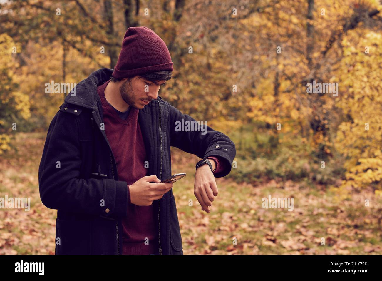 Kaukasischer weißer junger Mann synchronisiert Smartphone mit Smartwatch im Wald, gekleidet in langen Mantel und Mütze. Herbstfarben Stockfoto