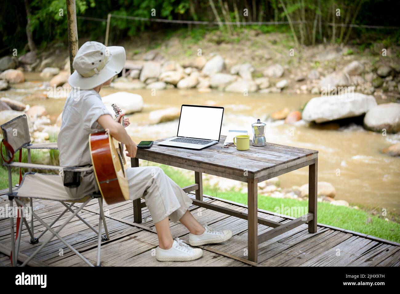 Entspannte junge asiatische Frau singt ein Lied, während sie Gitarre spielt und den Blick auf den wunderschönen natürlichen Fluss blickt. Campingkonzept Stockfoto