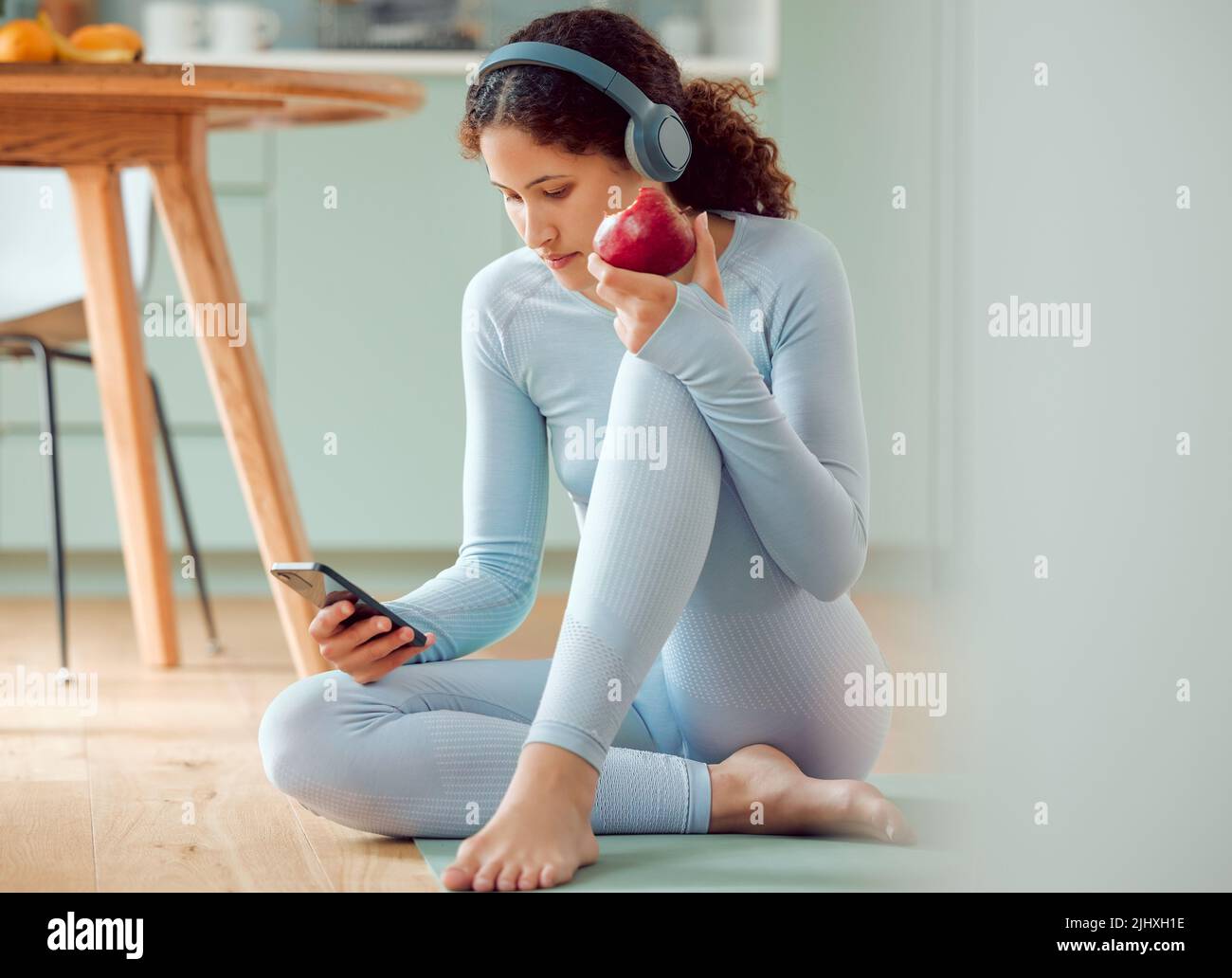 Schöne junge Frau mit gemischter Rasse, die Musik hört, während sie zu Hause eine Pause vom Yoga macht. Hispanische Frau, die ihr Telefon benutzt und eine isst Stockfoto