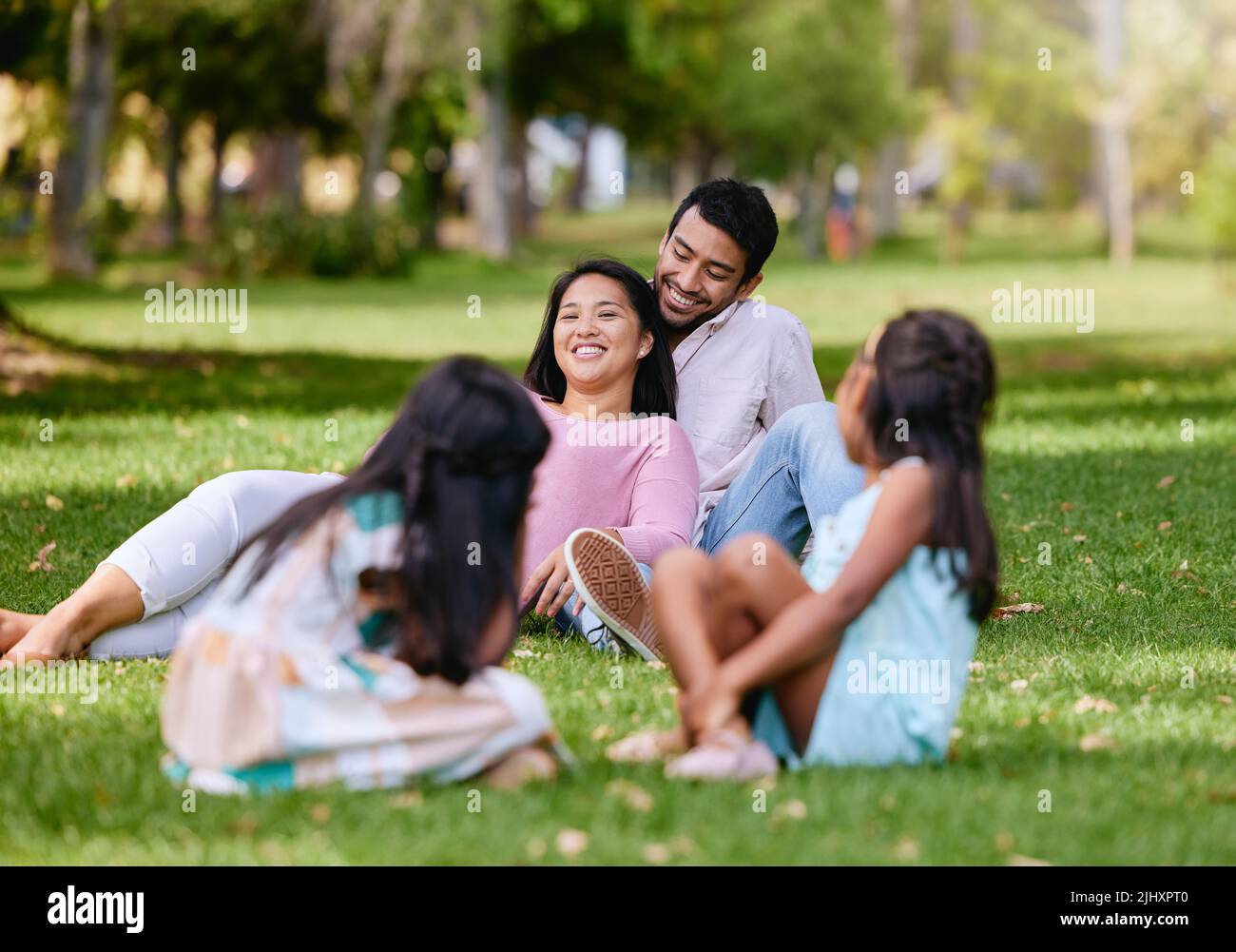 Portrait eines glücklichen asiatischen Paares, das auf Gras liegt. Kinder spielen, während sie ihren liebenden Eltern dabei zusehen, wie sie gemeinsam im Park Zeit verbringen Stockfoto