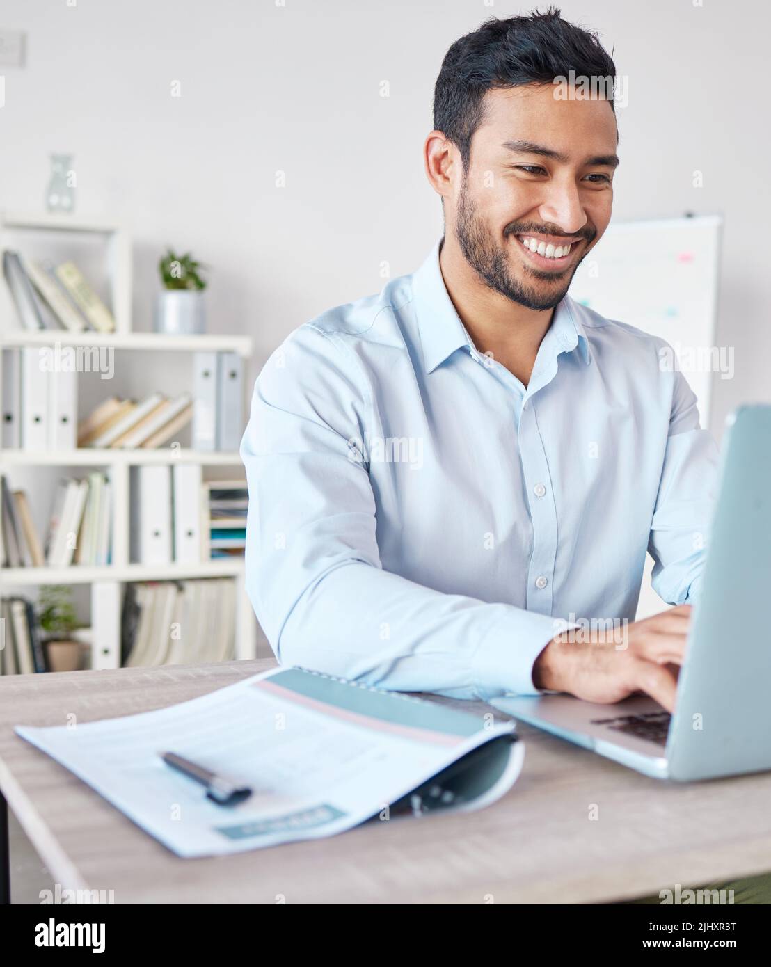 Junger glücklicher Geschäftsmann mit gemischtem Rennen, der allein in einem Büro auf der Arbeit an einem Laptop arbeitet. Ein hispanischer männlicher Chef lächelt, während er eine E-Mail tippt und eine verwendet Stockfoto