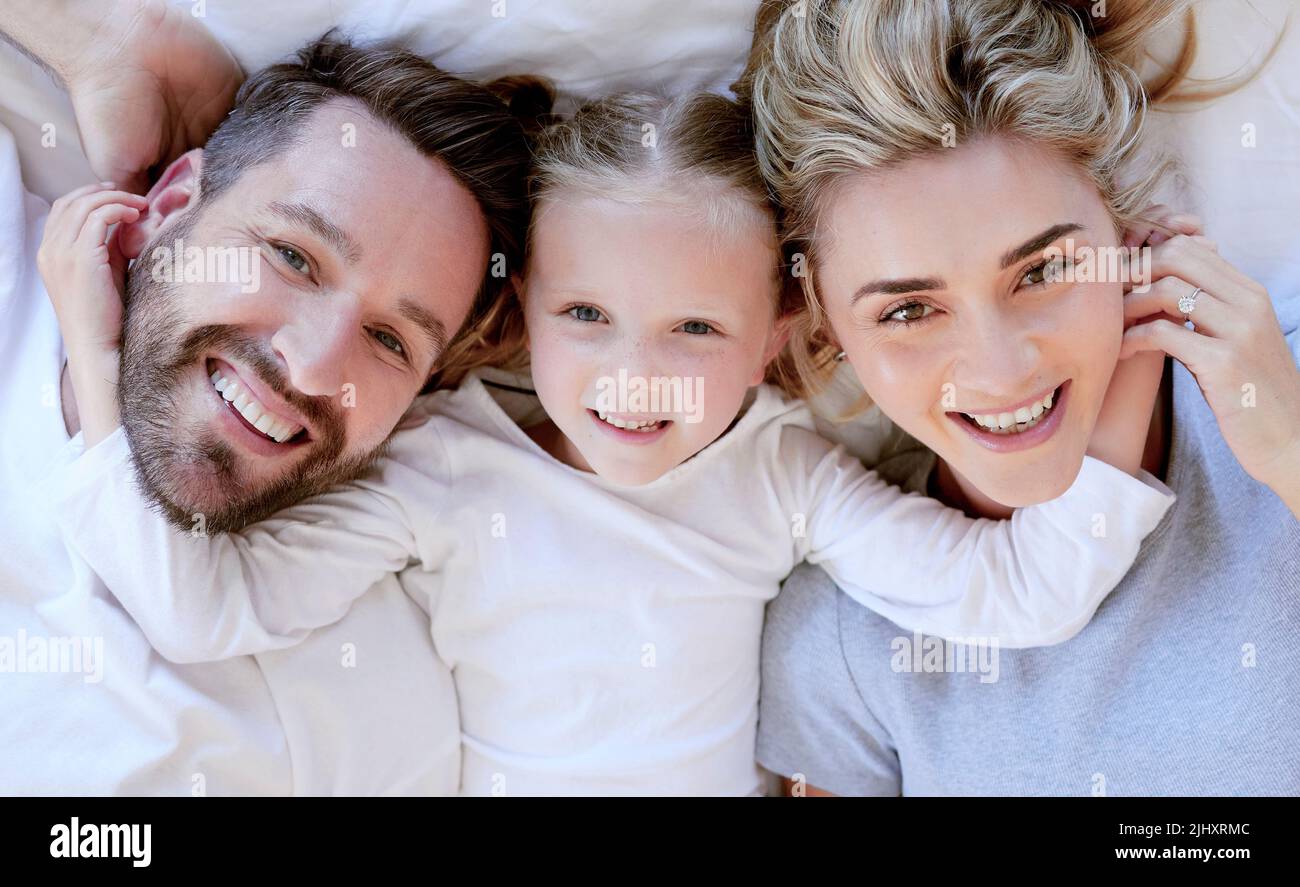 Porträt einer glücklichen, unbeschwerten kaukasischen Familie im Schlafanzug von oben, gemütlich im Bett zu Hause liegend. Liebevolle Eltern mit kleiner Tochter. Niedliche junge Leute Stockfoto