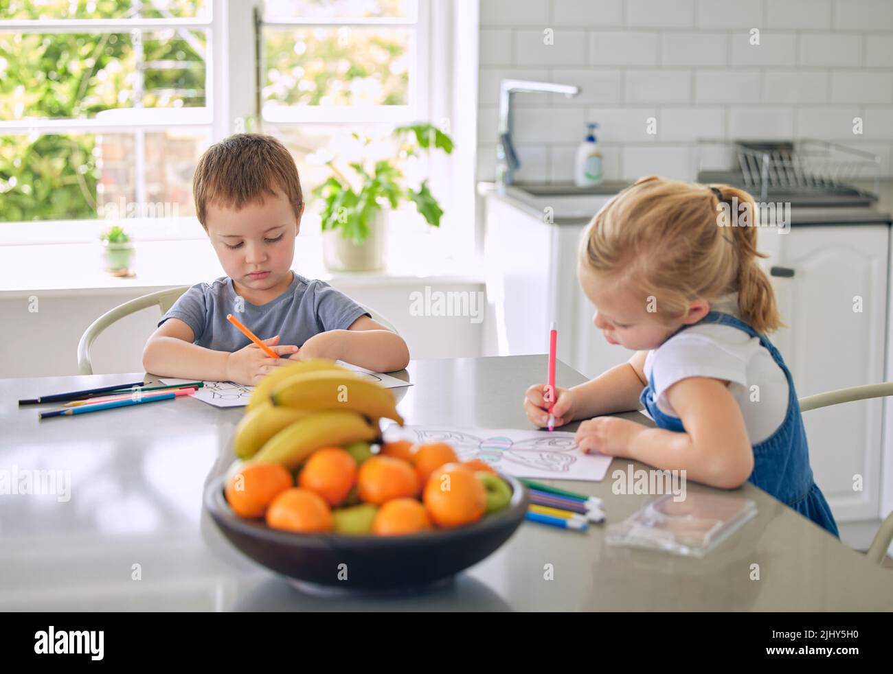 Kaukasische Geschwister zeichnen zusammen Bilder. Bruder und Schwester machen Hausaufgaben. Kinder färben in Skizzen in ihrer Küche. Kleines Mädchen Zeichnung Stockfoto