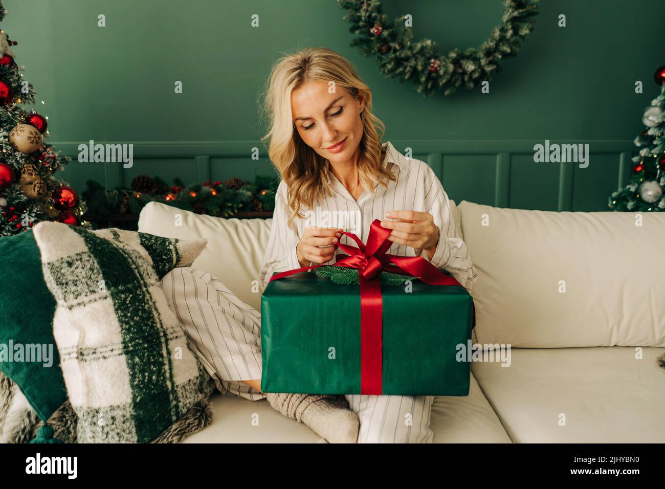 Eine junge Frau im Schlafanzug sitzt auf der Couch und wickelt ein Weihnachtsgeschenk aus. Stockfoto