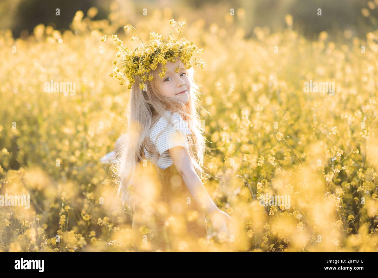 Nette stilvolle Kind Mädchen 4-5 Jahre alt tragen Blumenkranz stehen in gelben Blumenwiese im Freien über Natur Hintergrund über Sonnenlicht. Kindheit. Sommer Stockfoto