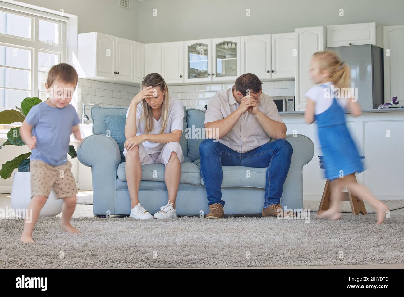 Müde und gestresste Eltern, die unter Kopfschmerzen leiden, während adhd-Kinder im Wohnzimmer herumlaufen. Laute freche Geschwister ärgern ihre Mutter und Stockfoto