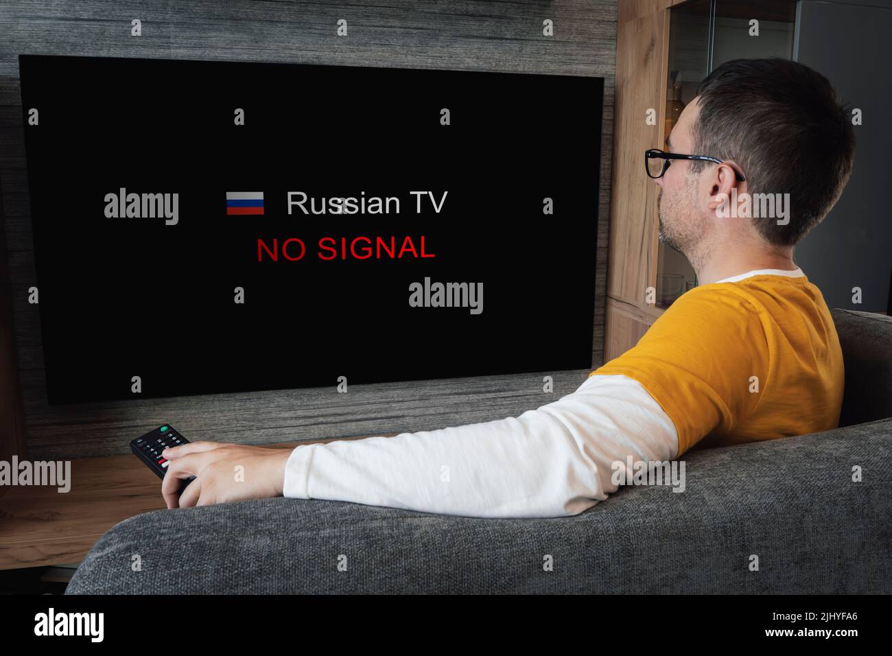 Das Konzept der Trennung russischer Sender in Europa und den USA. Sperrung der russischen Fernsehübertragung. Junger, hübscher Mann, der auf einem Sofa Fernsehen sieht Stockfoto