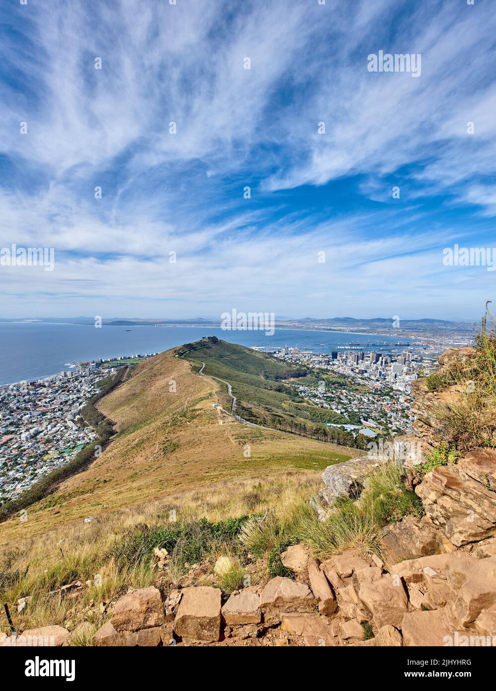 Felsiger Berg mit Blick auf eine Küstenstadt am Meer in Kapstadt. Landschaft von grünen Hügel mit Steinen und Pflanzen umgeben von einer Stadt und Stockfoto