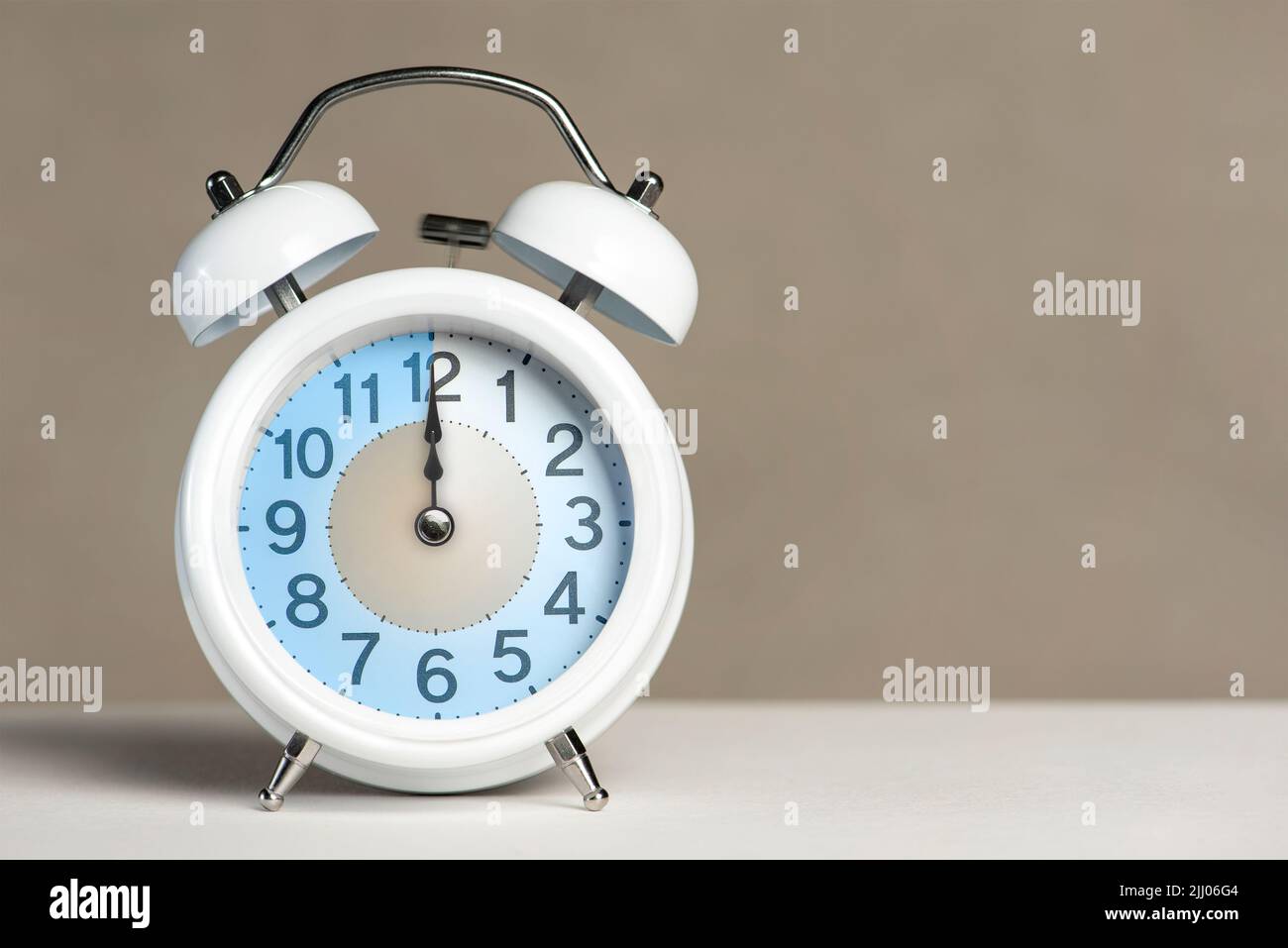 Zwölf Uhr auf dem Wecker. Ein weißer Wecker befindet sich auf einem weißen Tisch. Der Uhrzeiger zeigt auf 12 Uhr. Zeit, um in Sommer- oder Winterzeit zu wechseln Stockfoto