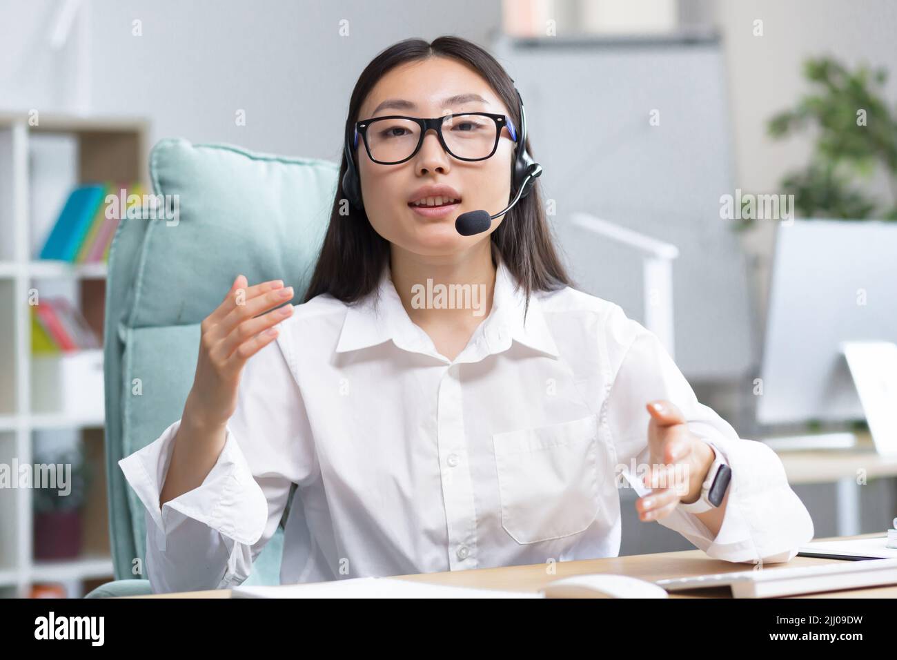Online-Fernunterricht. Junge schöne asiatische Lehrerin mit Kopfhörern und Brille sitzt an einem Tisch an einem Computer und unterrichtet online eine Klassenstunde. Stockfoto