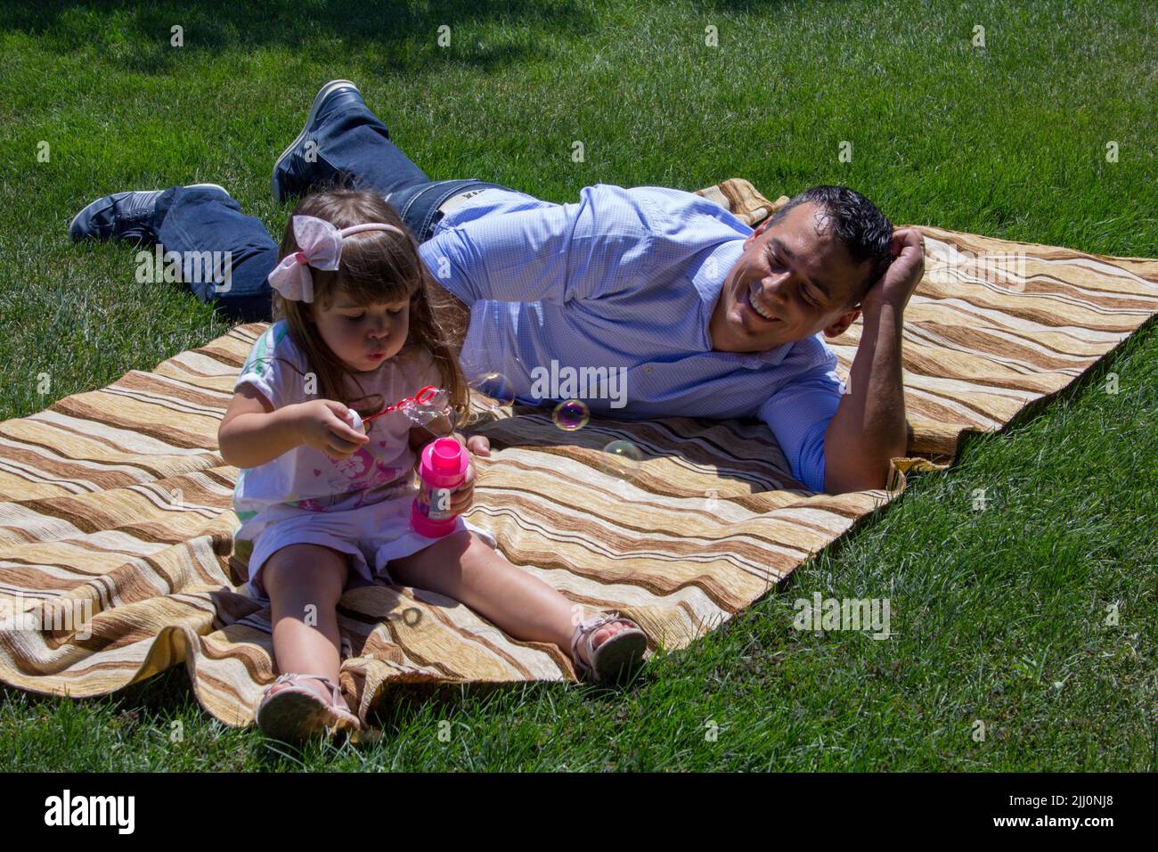 Bild eines jungen Vaters, der auf einer grünen Wiese sitzt und mit seiner Tochter spielt, die Seifenblasen macht. Bezug auf die Bindung zwischen Vater und Tochter Stockfoto