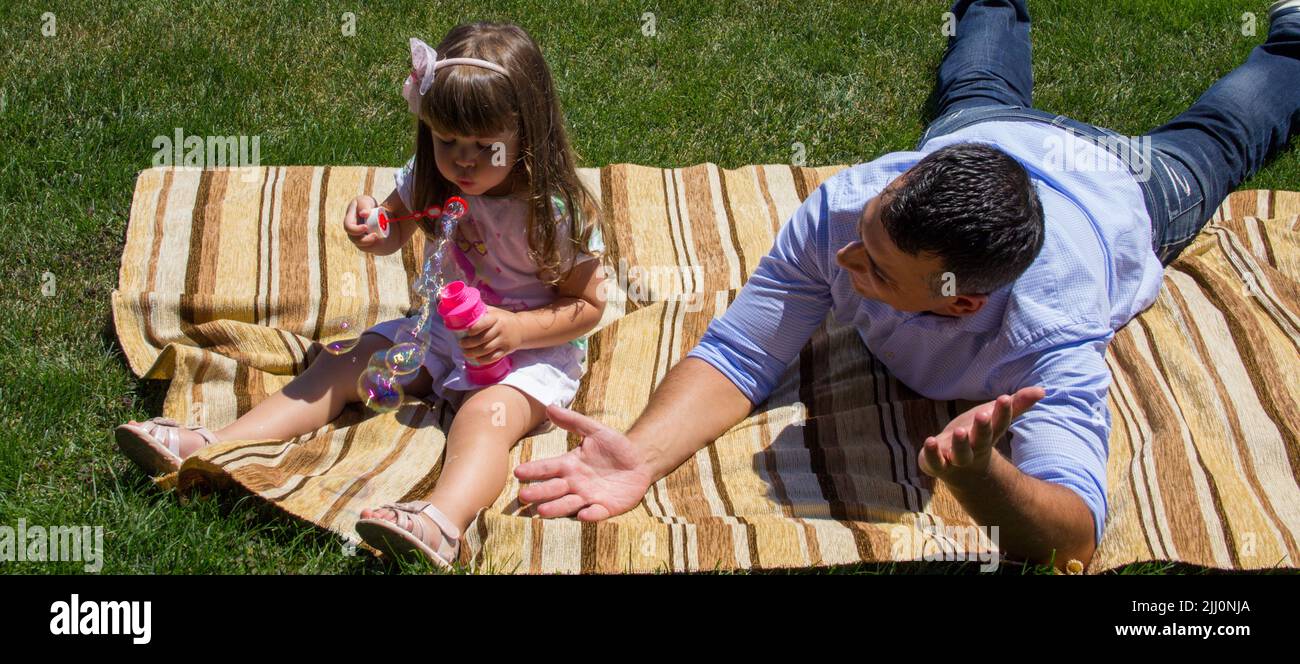 Bild eines jungen Vaters, der auf einer grünen Wiese sitzt und mit seiner Tochter spielt, die Seifenblasen macht. Hinweis auf die Bindung zwischen Vater und Tochter. Banner Stockfoto