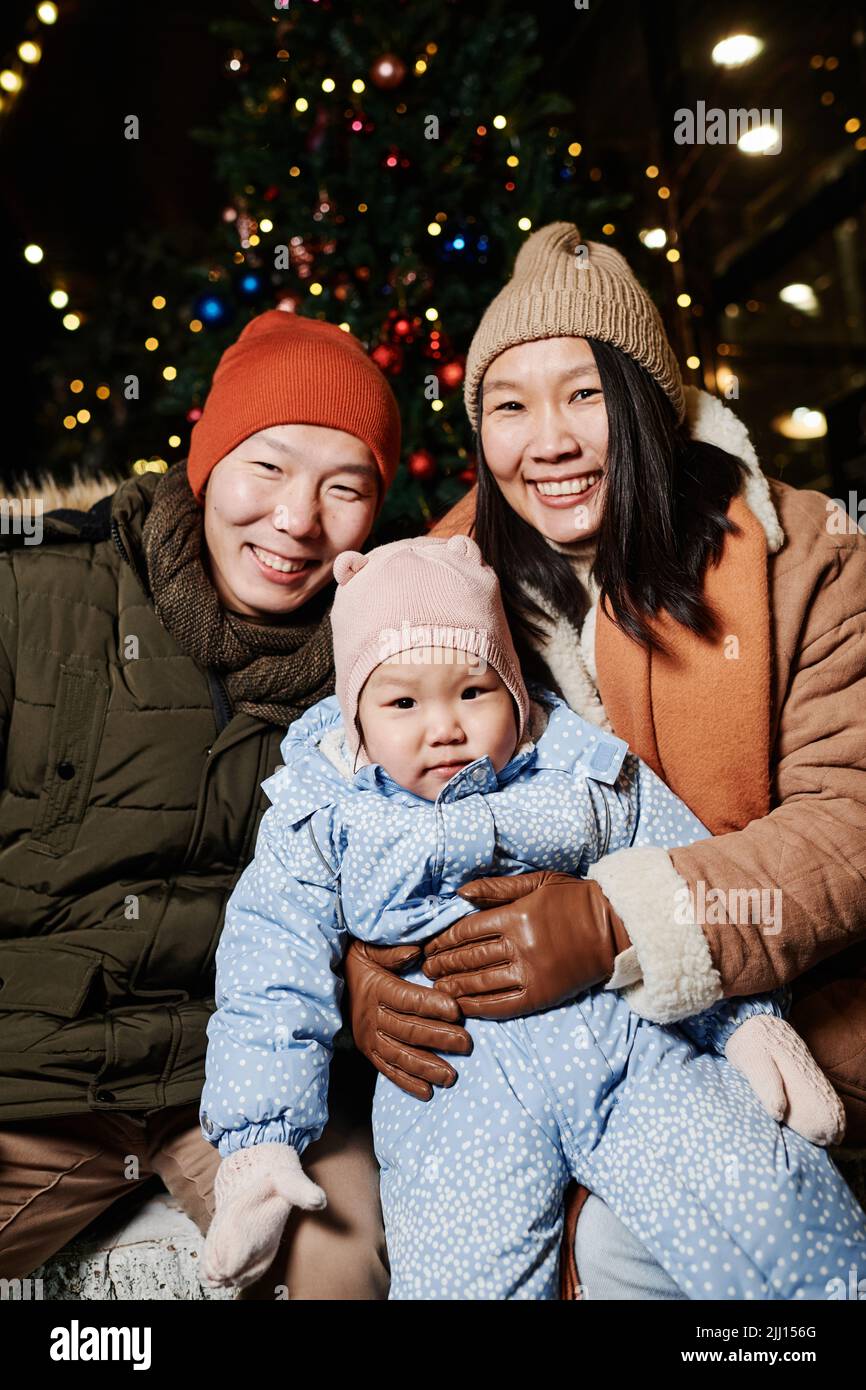 Vertikale mittlere Aufnahme von fröhlichen asiatischen Eltern mit ihrer hübschen Kleinkindtochter, die am Winterabend Zeit im Freien verbringt und lächelt vor der Kamera Stockfoto