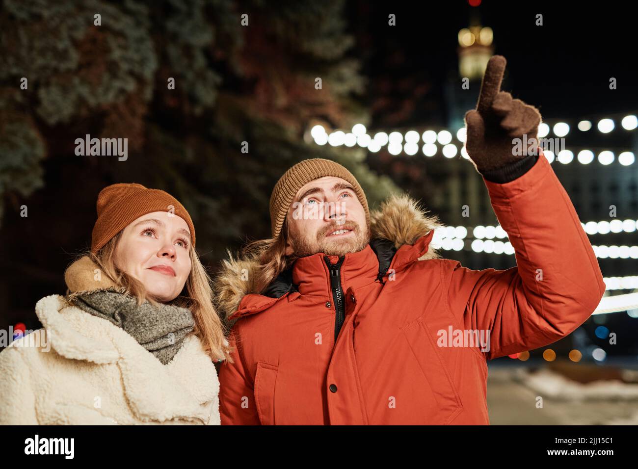 Horizontale Aufnahme eines jungen Paares, das am Winterabend spazierengeht, wobei der Mann seiner Freundin etwas zeigt Stockfoto