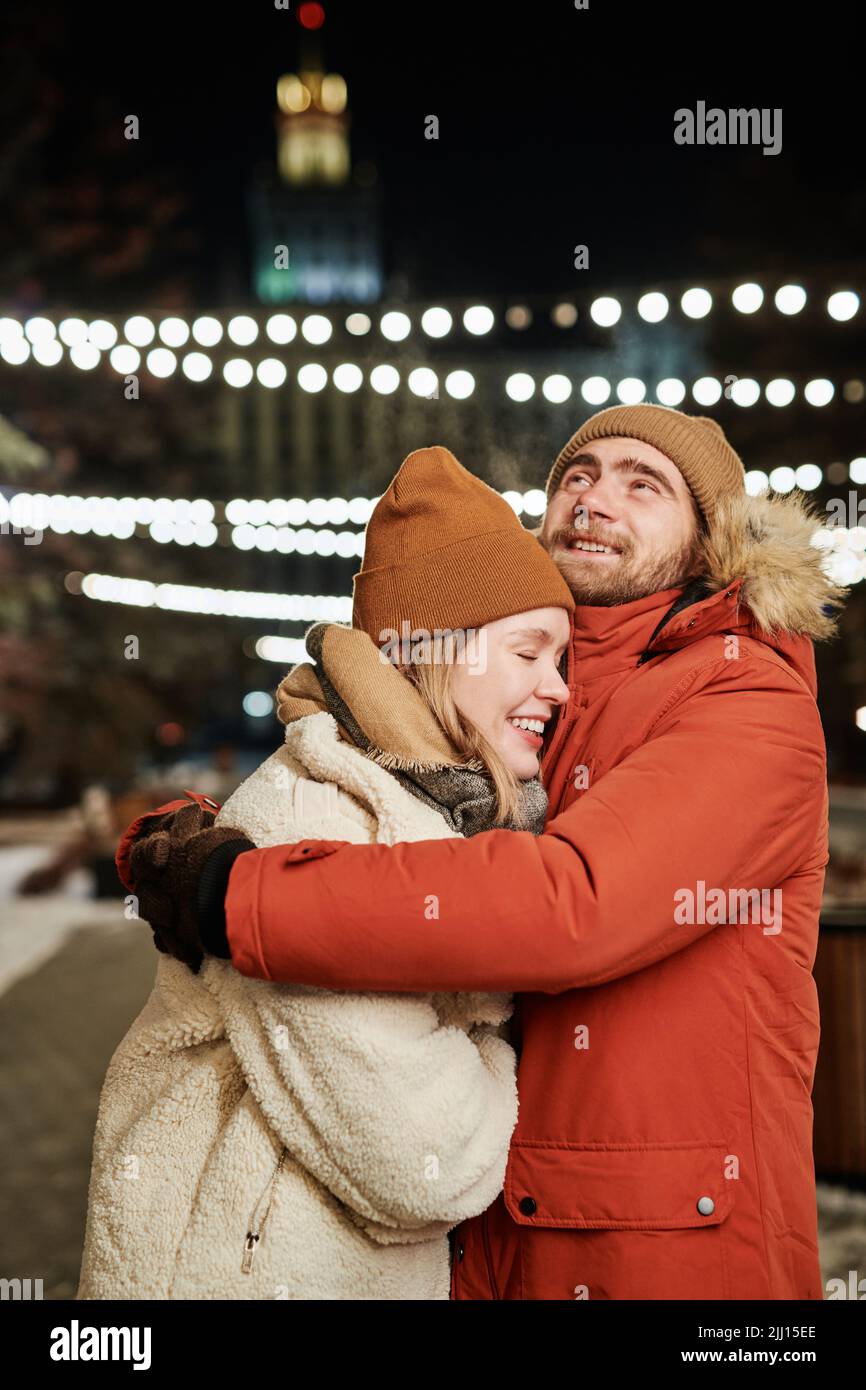 Vertikale Aufnahme eines glücklichen jungen Paares, das den Winterabend im Freien verbringt, umarmt und lächelt Stockfoto