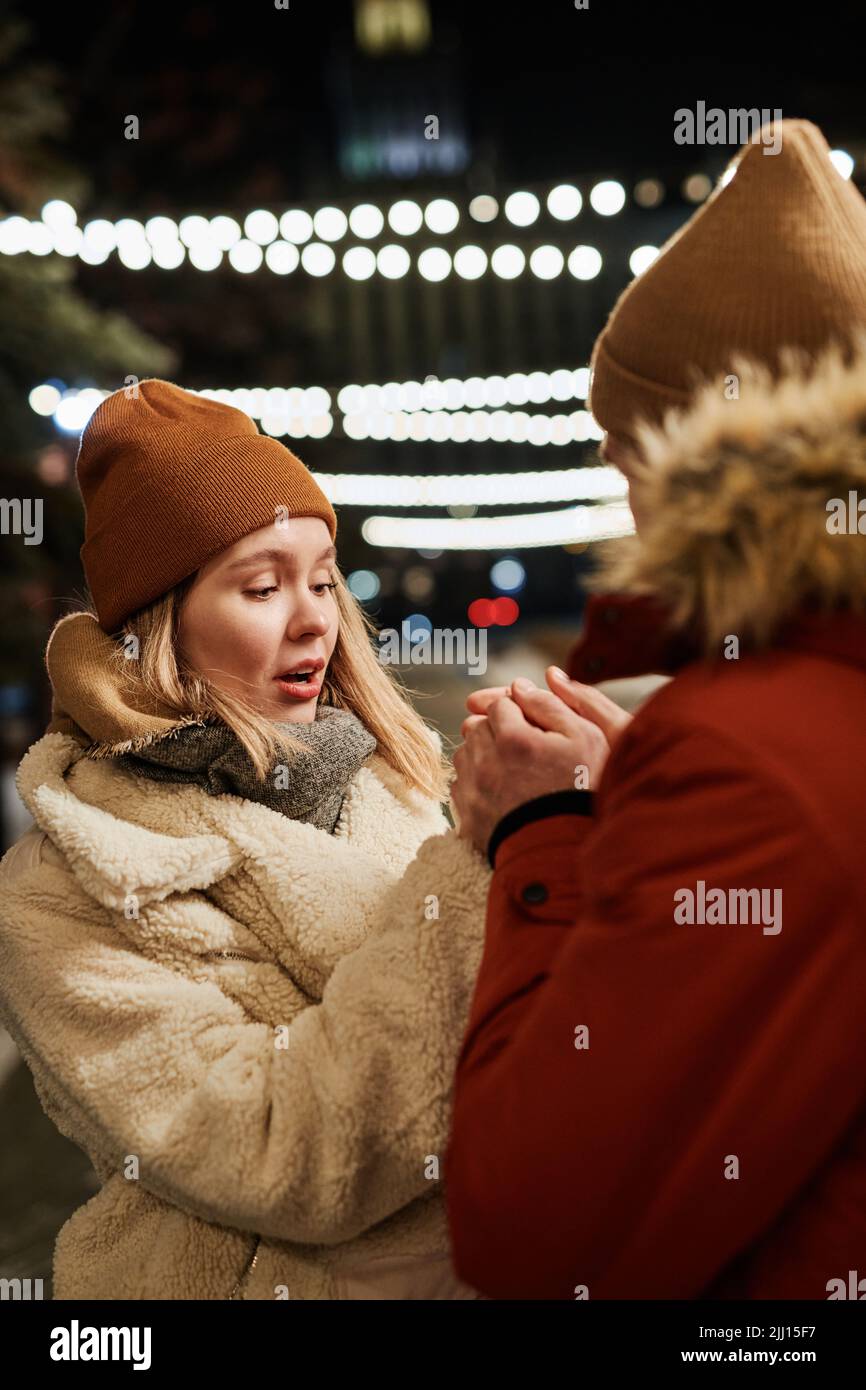 Vertikale mittlere Aufnahme eines verliebten jungen Mannes, der am Winterabend im Freien steht und seine Freundin sich die Hände wärmt Stockfoto