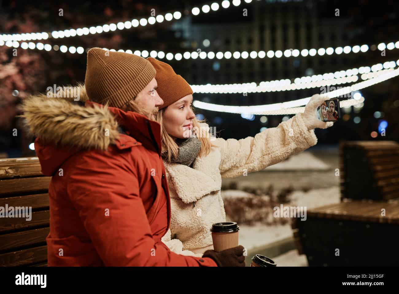 Horizontale mittlere Aufnahme eines modernen jungen Paares, das den Winterabend auf einer Bank im Freien verbringt und Selfies auf einer Smartphone-Kamera macht Stockfoto