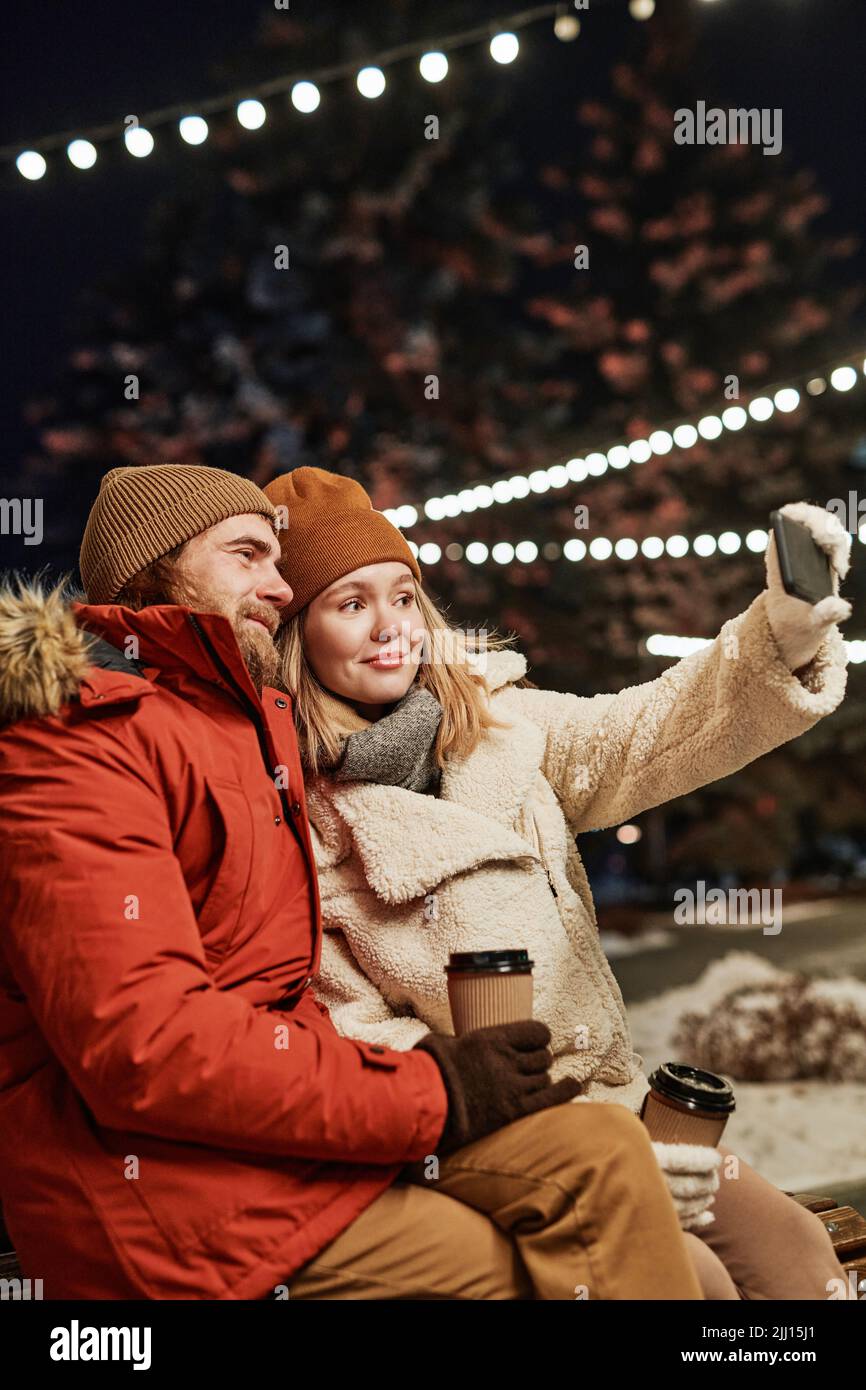 Vertikale mittlere Aufnahme des jungen kaukasischen Paares verbringen Winterabend sitzen auf der Bank im Freien unter Selfies auf Smartphone-Kamera Stockfoto
