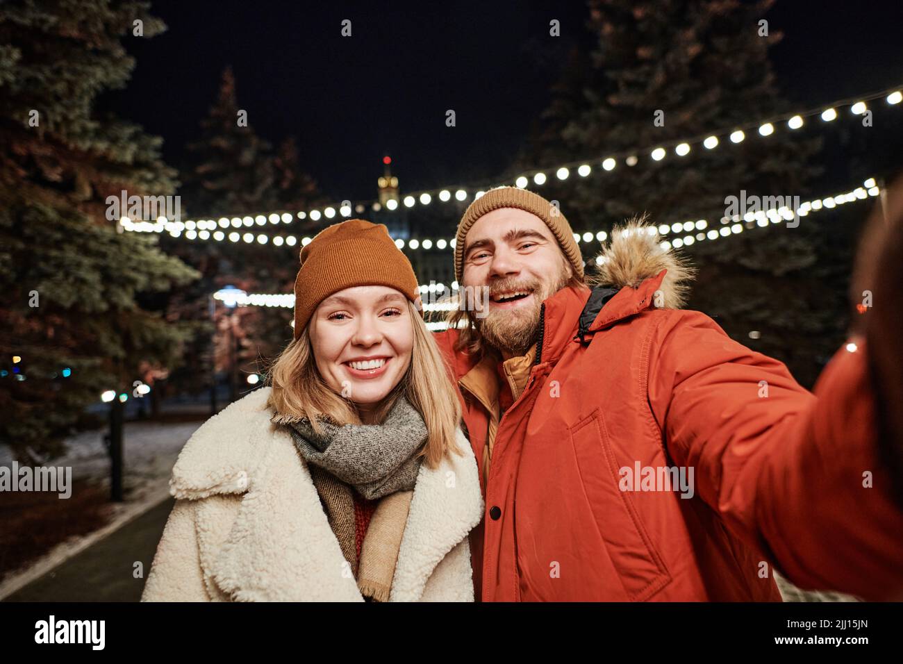 Horizontal Medium shot fröhliches junges Paar, das Winterabend im Freien verbringt und Spaß dabei hat, Selfies auf der Smartphone-Kamera zu machen Stockfoto