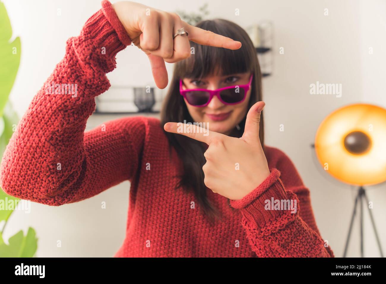 Kaukasische Frau mit dunklem Haar trägt eine rosa Sonnenbrille und einen roten Pullover, der das Gesicht mit den Händen umrahmt. Innenaufnahme. Hochwertige Fotos Stockfoto