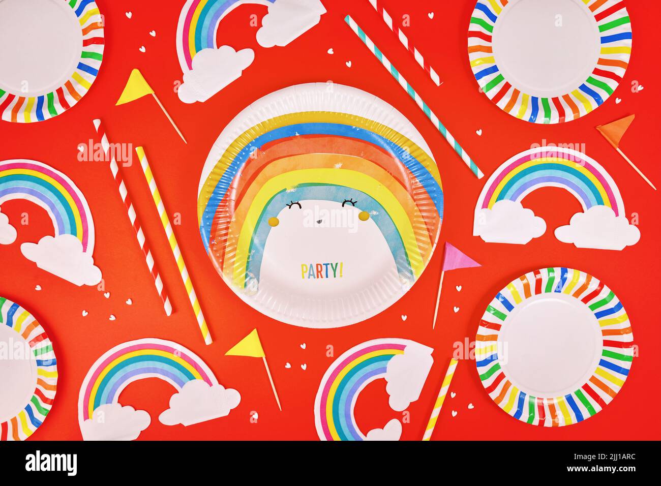 Party Flat Lay mit bunten Tellern, Regenbogenservietten und Trinkhalmen auf rotem Hintergrund Stockfoto