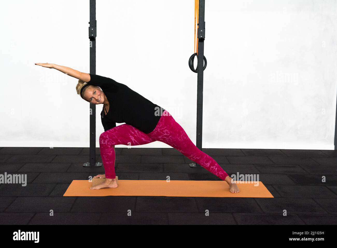 Eine blonde Jugenddame in einem schwarzen Hemd und pinken Leggings, die in einem Fitnessstudio die Utthita Parshvakonasana-Yoga-Pose mit erweitertem Seitenwinkel ausführt. Stockfoto