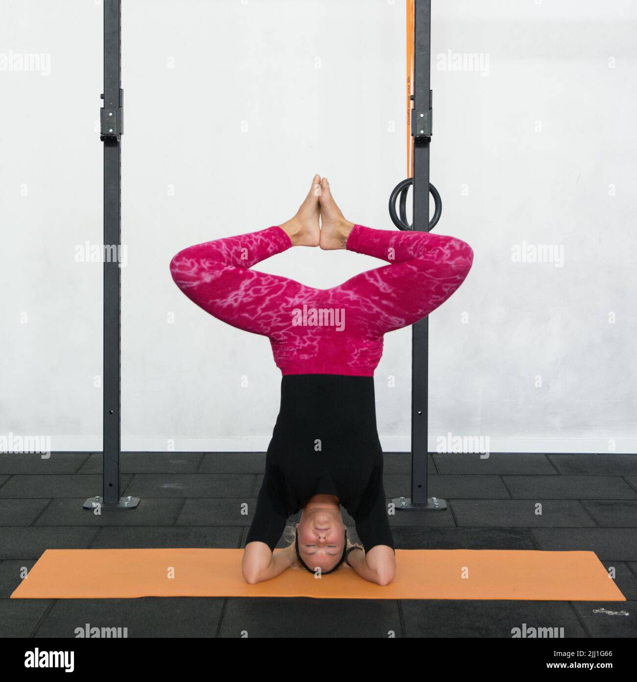 Blonde junge Frau, die Kopfstand mit gebeugten Knien und Fuß zusammen Yoga-Pose in rosa Yoga-Leggings und schwarzem Hemd in einem Fitnessstudio. Stockfoto