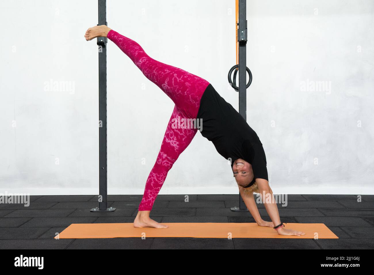 Eine blonde junge Frau führt die Dog Facing Down Pose mit dem linken Bein nach oben aus. Lächelnd zur Kamera. Fotoshooting im Fitnessstudio. Rosa Yoga Legging. Stockfoto