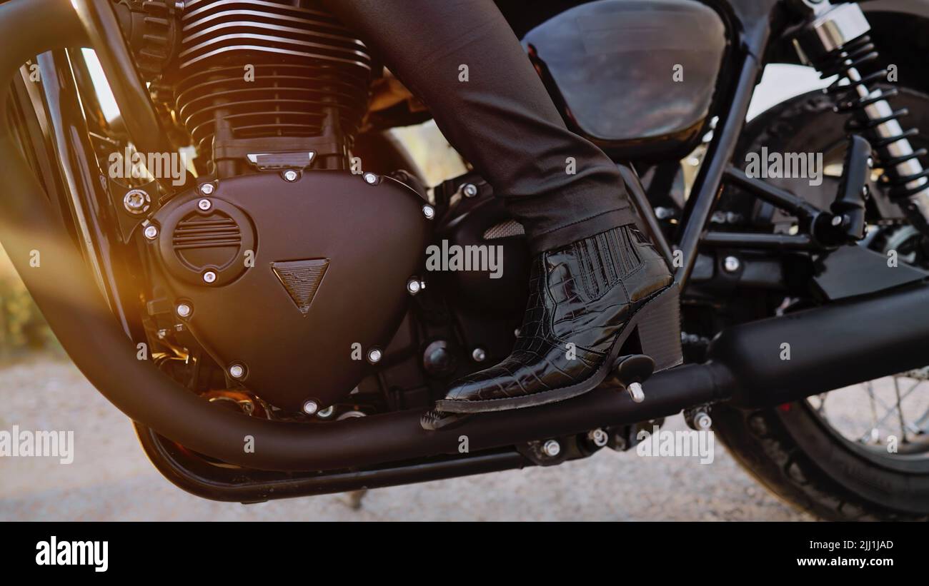 Die Beine der stilvollen Motorradfahrerin Frau, die auf einem klassischen Fahrrad sitzt. Schwarzes Motorrad im Retro-Stil. Details des Vintage-Designs des brandneuen Motorrads. Sonneneinstrahlung Stockfoto