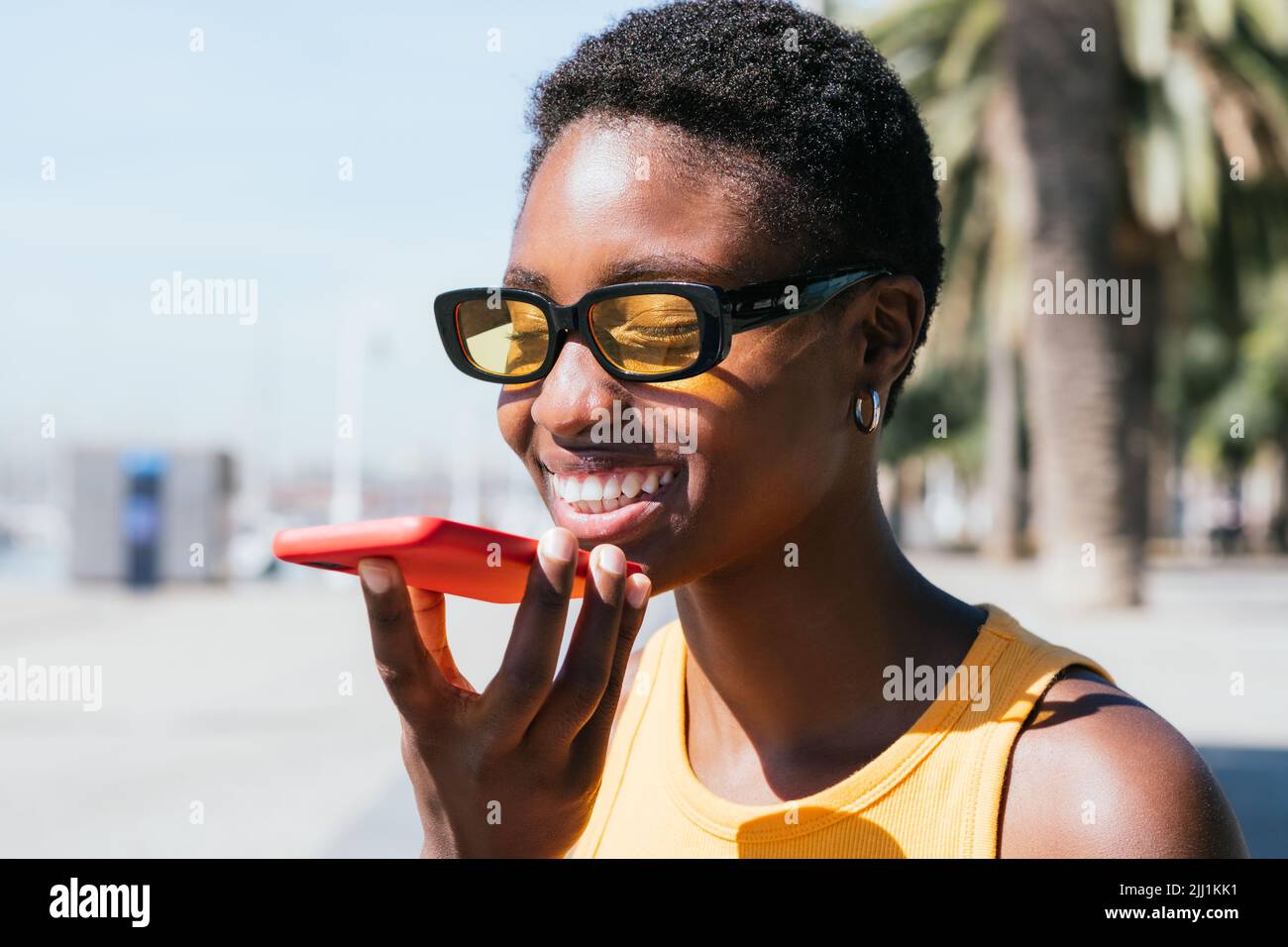 Fröhliche afrikanische Frau, die an einem sonnigen Tag eine Sprachnachricht im Freien sendet. Sie trägt lässige Sommerkleider und lächelt fröhlich Stockfoto