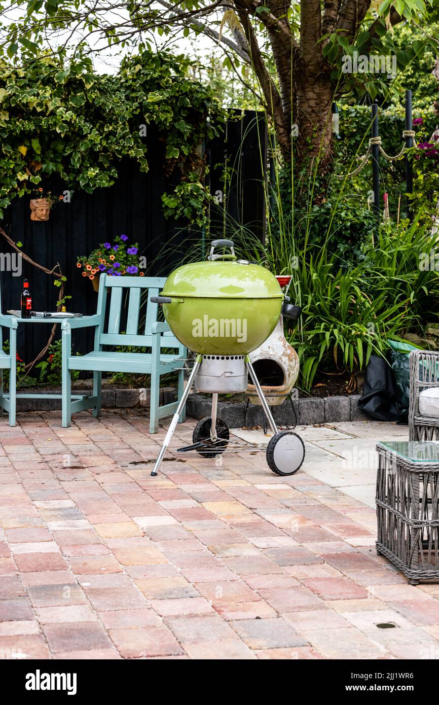 Grüner Wasserkocher Grill auf einer Stein/Fliesen Terrasse, mit Stuhl und Tisch mit einer Bierflasche im Hintergrund, mit Gartensträuchern und Holztäfelung Stockfoto