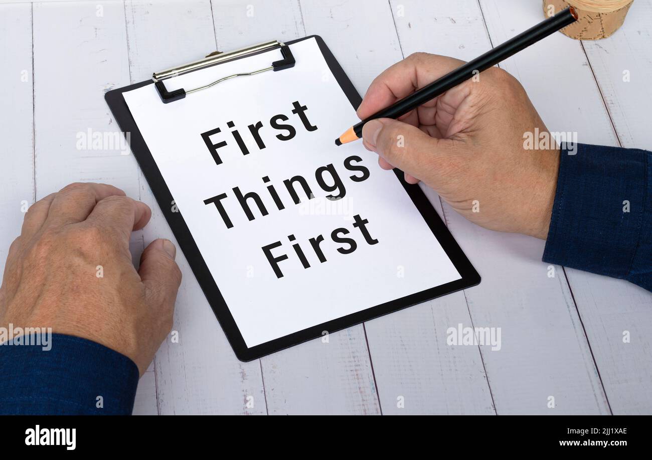 Hand mit Bleistift, der die Wörter zuerst auf ein Papier mit schwarzer Zwischenablage schreibt, um Sie an eine wichtige Aufgabe oder einen wichtigen Termin zu erinnern. Stockfoto