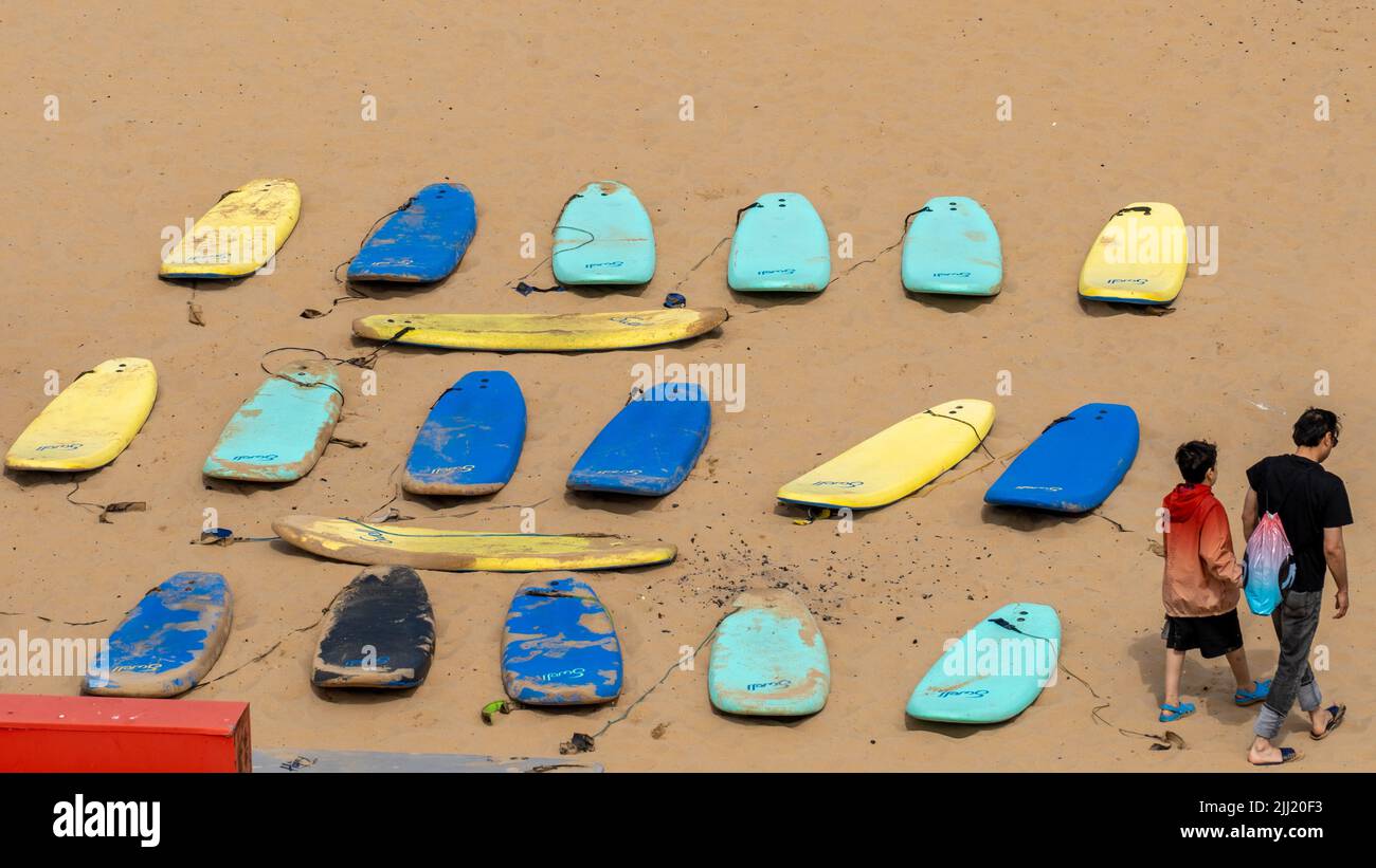 Reihen von Surfbrettern mit zwei Surfern, die am Long Sands Beach, Tynemouth, North Tyneside, Großbritannien vorbeilaufen Stockfoto