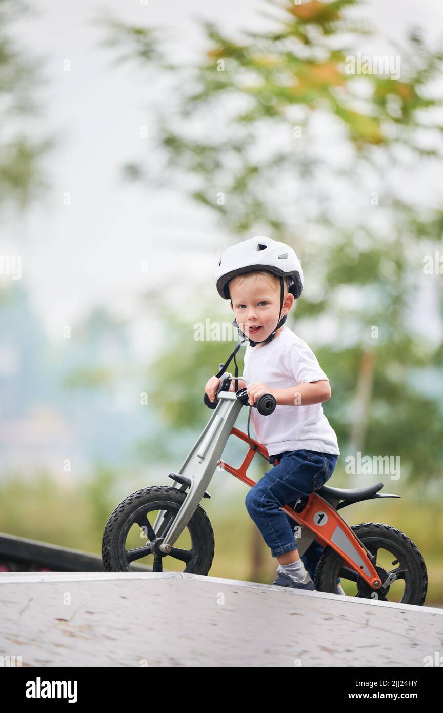 Porträt des Kindes Reiten Laufrad. Männliches Kleinkind im Helm, das im Skatepark lernt, auf dem Laufrad zu fahren. Stockfoto