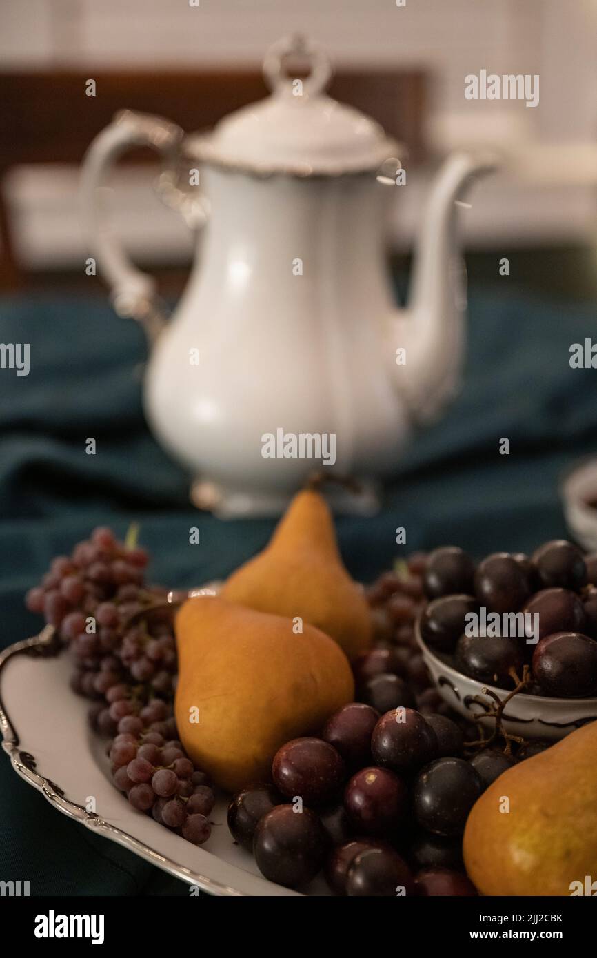 Auf einer dunkelgrünen Tischdecke vor einer teekannen aus china sitzt eine weiß-silberne Platte aus violetten Muscadinen, winzigen Trauben und Bosc-Birnen. Stockfoto