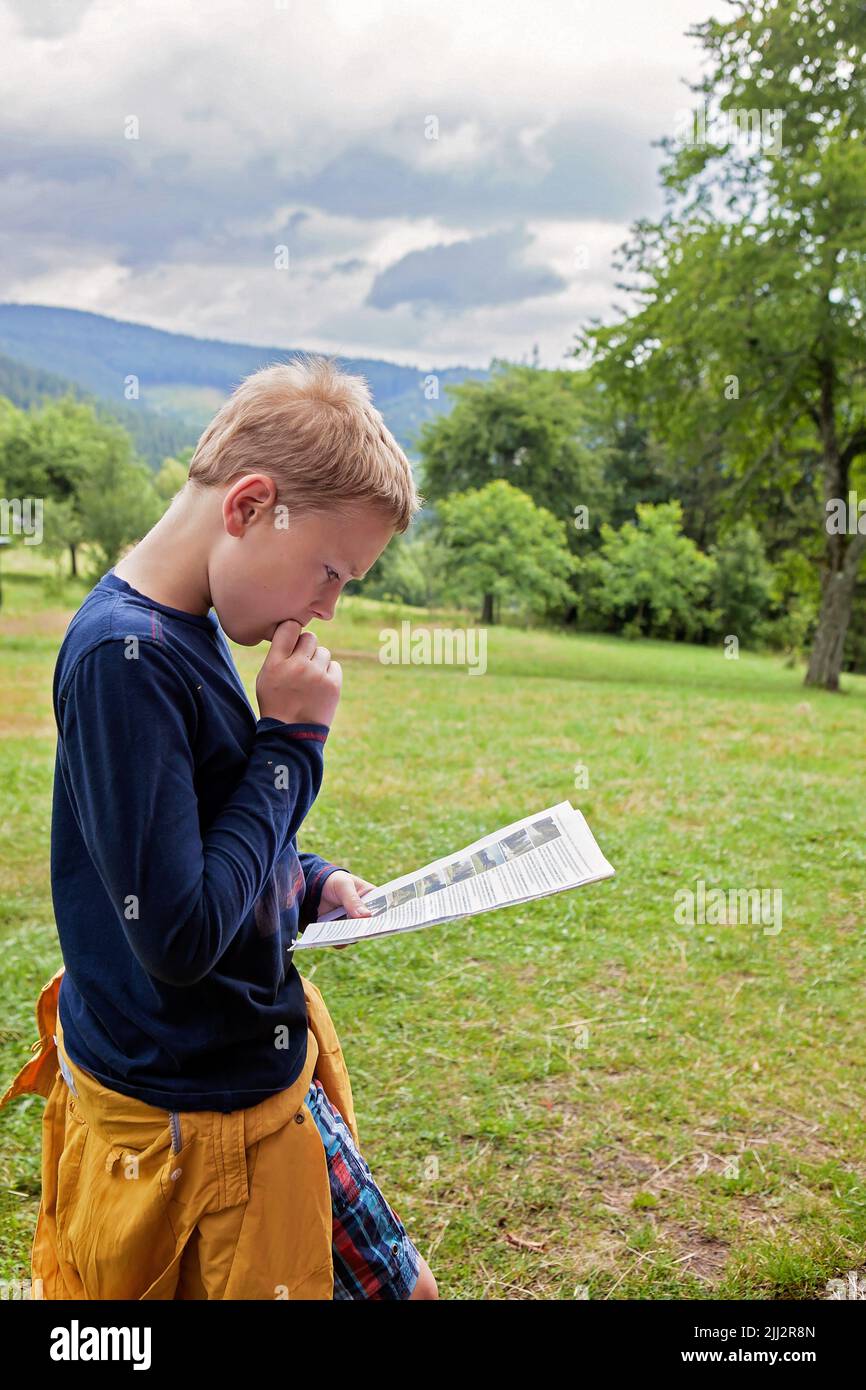 Kleiner Junge, der eine Karte, ein Papier, Anweisungen in einem Berghochland, Wald hinter sich liest. Auf einer Wiese, Besuch, Urlaubsreise, tourisme. Zehn Jahre alt Stockfoto