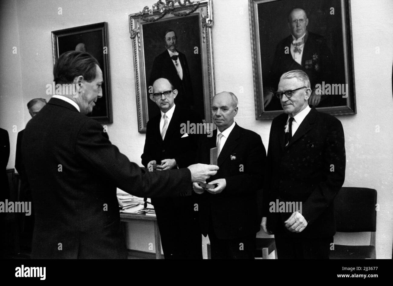 Örebro Skofabrik schließt, der bezirksrat teilt Auszeichnung, Weihnachtsbäume 19. Dezember 1967 Herr in schwarzem Anzug steht mit dem Rücken gegen die Kamera und hält ein Ding in seiner rechten Hand, das er ausgestreckt hält. Im Hintergrund sind drei Herren in schwarzen Anzügen, weißen Hemden und hellen Krawatten zu sehen. Zwei von ihnen halten kleine Schmuckkisten in ihren Händen. An der Wand hinter ihnen hängen drei Gemälde. Stockfoto