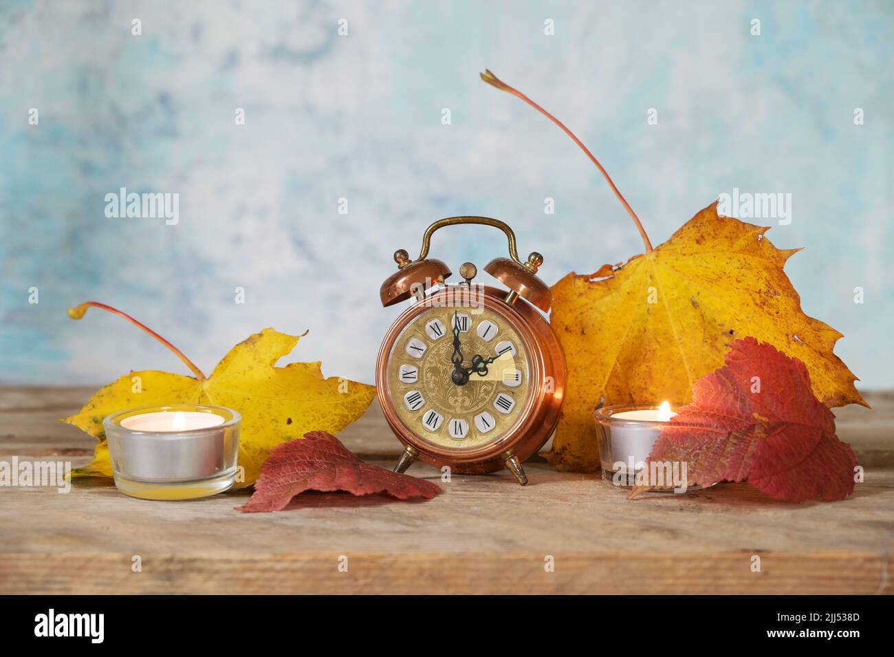 Wechsel von Sommer- auf Winterzeit, Vintage-Wecker mit einstündiger Rückstellung, Herbstblätter und Kerzen auf rustikalem Holz, hellblauer Hintergrund Stockfoto