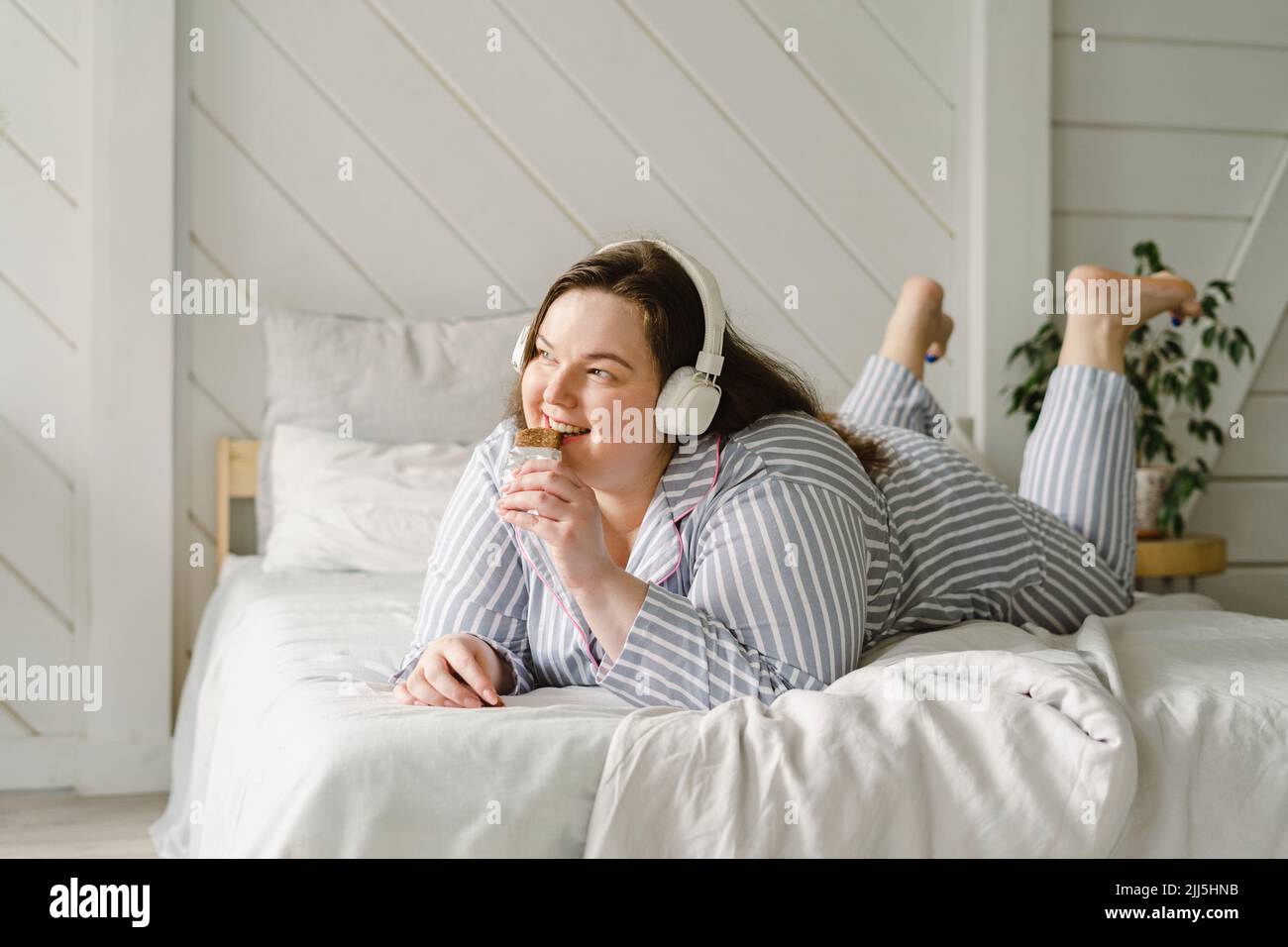 Glückliche Frau, die Schokolade isst und Musik über kabellose Kopfhörer hört, die zu Hause auf dem Bett liegen Stockfoto