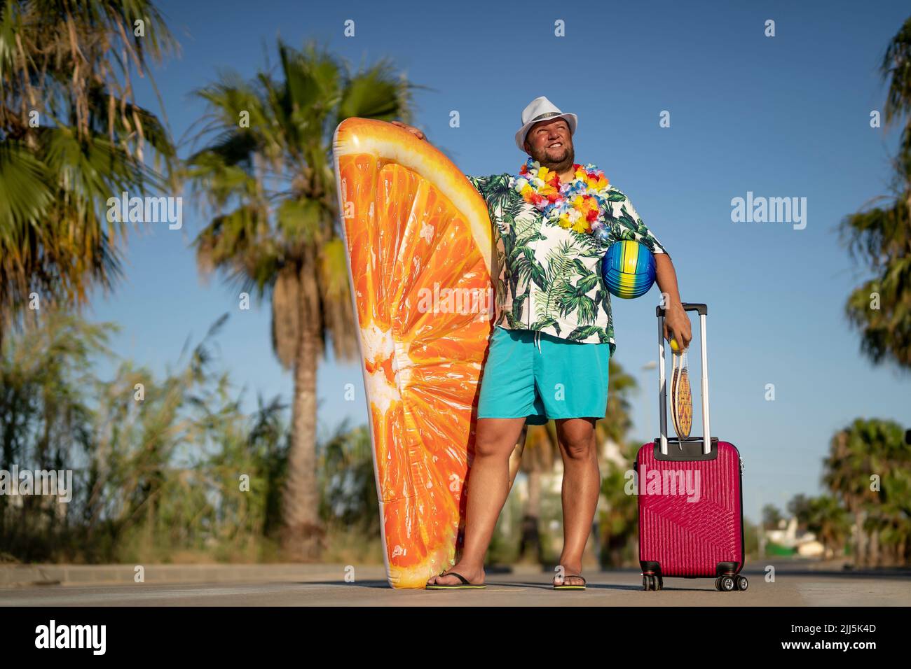 Lächelnder Mann, der ein orangefarbenes, in Scheiben gehaltenes Poolfloß und einen Koffer auf der Straße hält Stockfoto