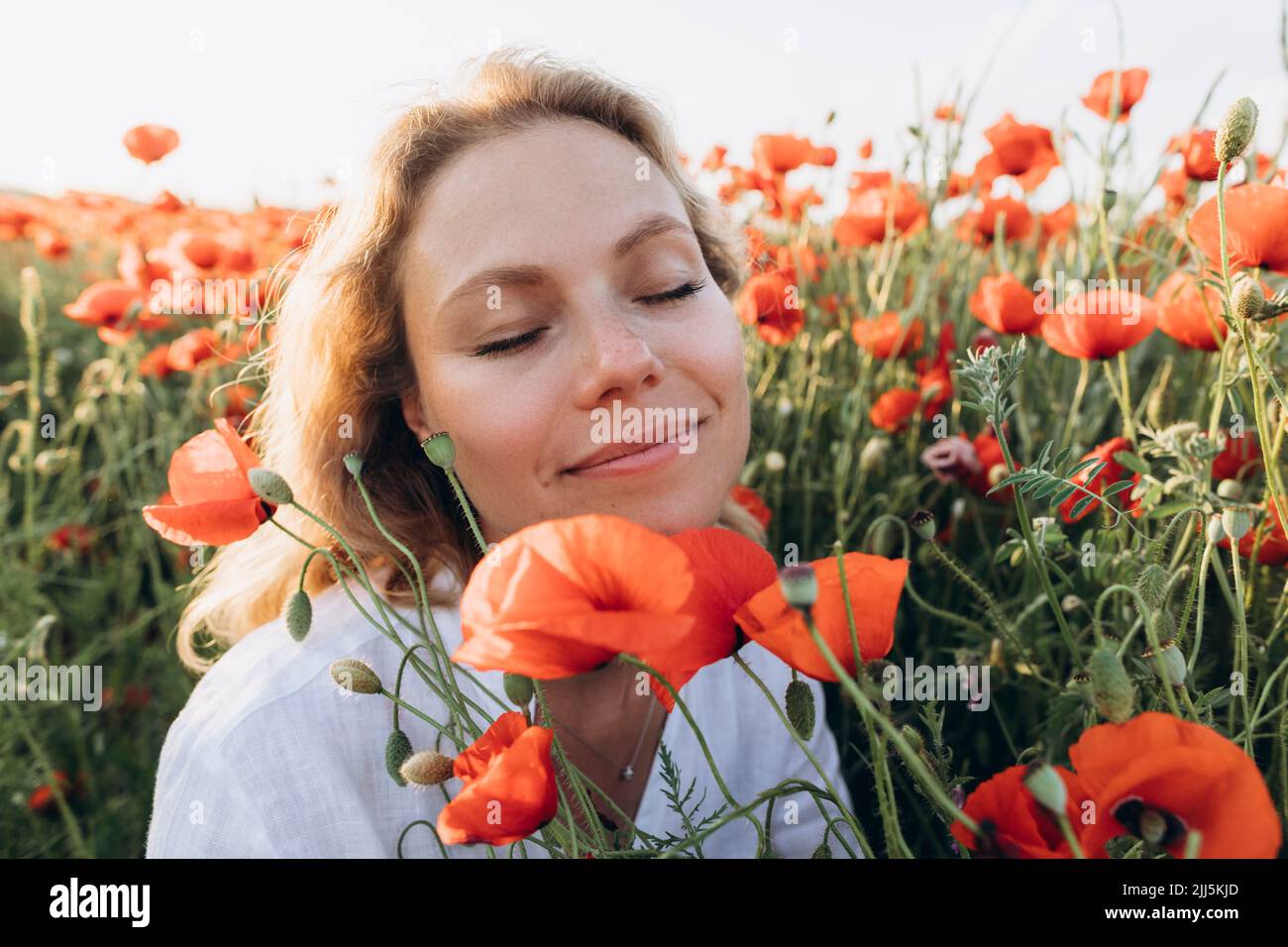 Lächelnde Frau mit geschlossenen Augen sitzt inmitten roter Blumen auf dem Feld Stockfoto