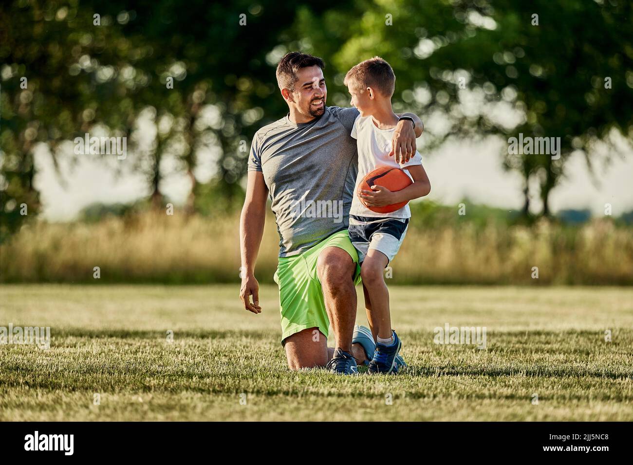 Junge hält Rugby-Ball und spricht mit Vater, der auf dem Sportplatz kniet Stockfoto
