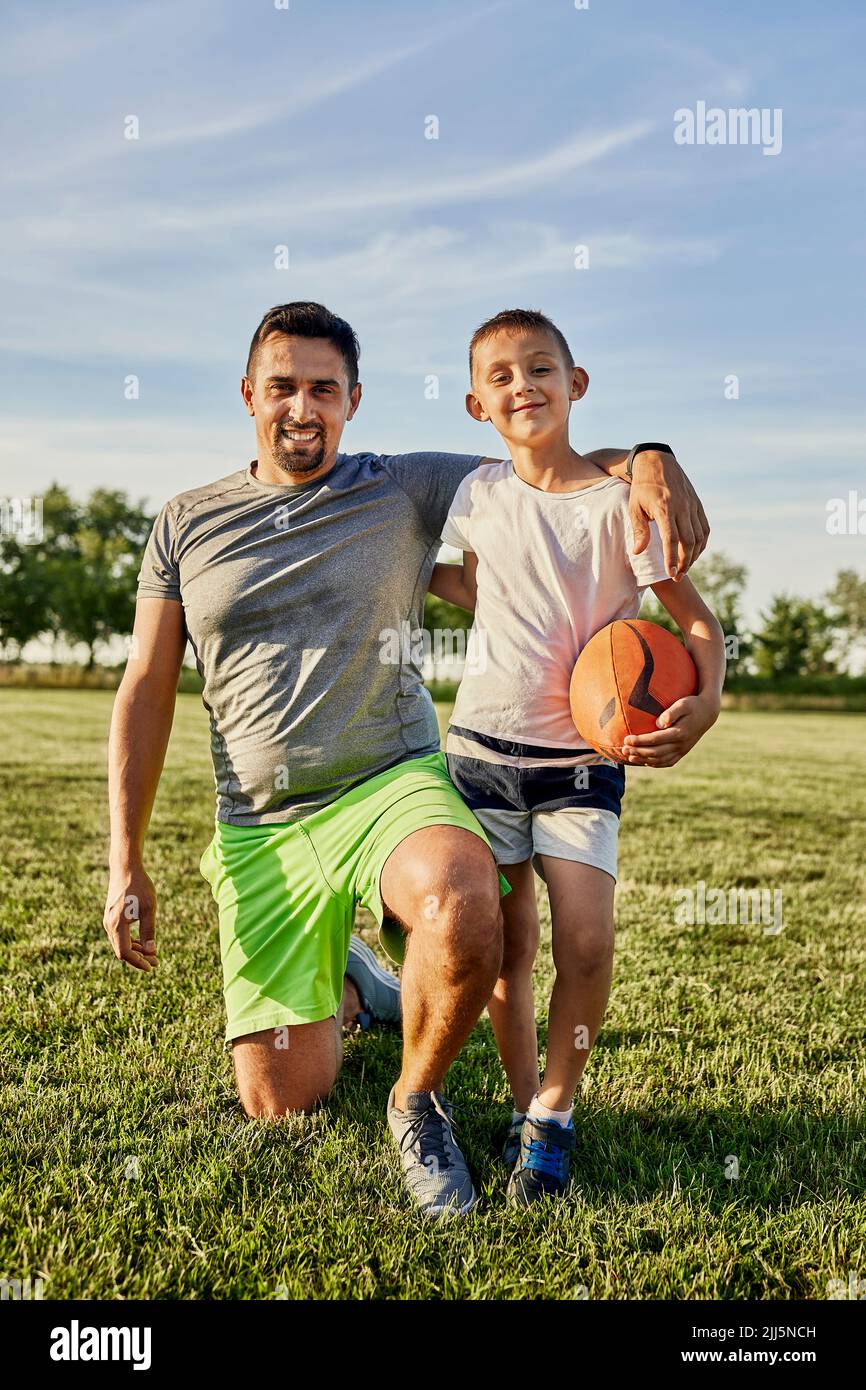 Glücklicher Mann kniet bei Sohn, der Rugby-Ball auf dem Sportplatz hält Stockfoto
