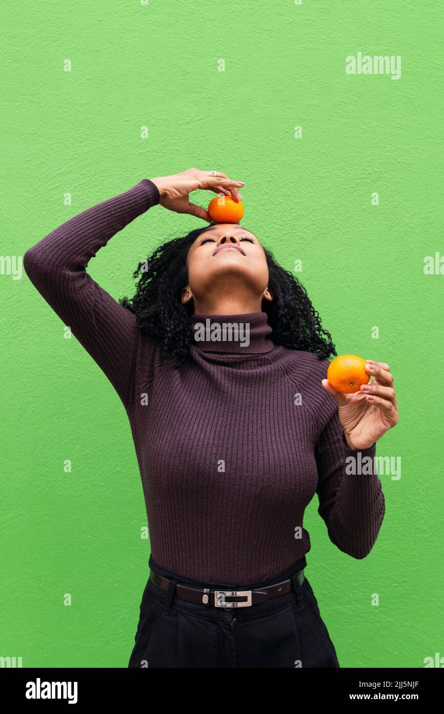Frau mit geschlossenen Augen balanciert orange auf dem Kopf vor der grünen Wand Stockfoto